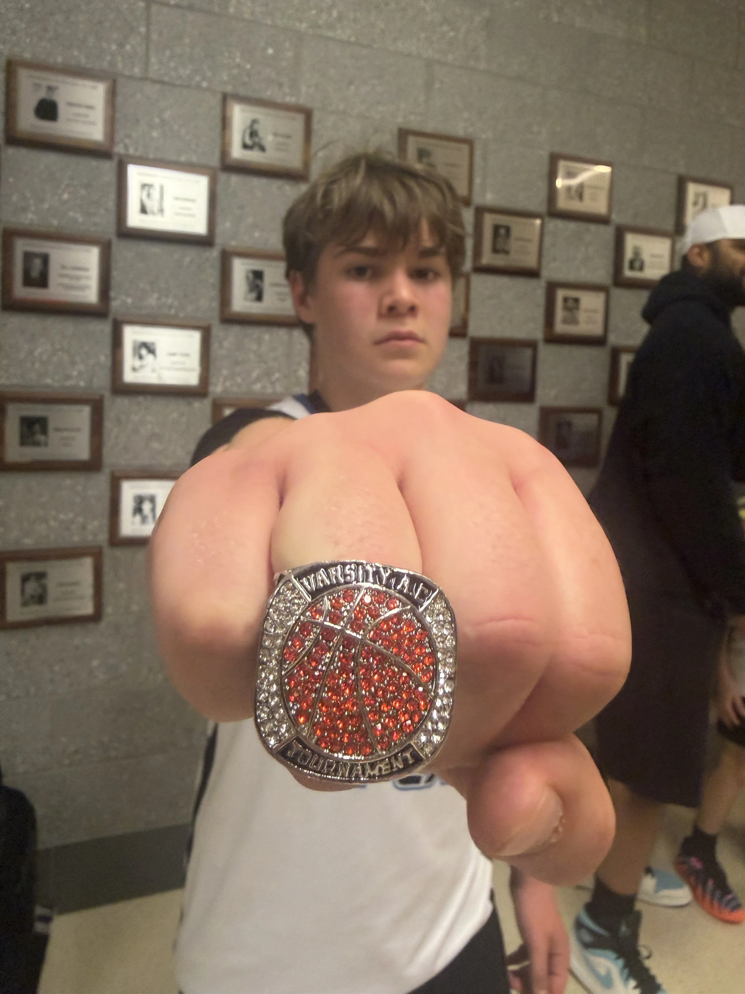 A young person is taking a selfie, showing a large championship belt in front of their chest. The belt has a red and orange design with rhinestones and the words 'WORLD CHAMPION' on it. The background features a wall with framed photographs or plaques, and another person is visible in the background wearing a white cap and black jacket.