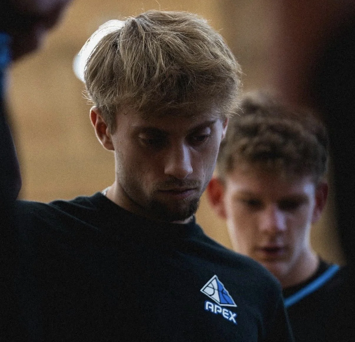 Two young men with blond hair are looking down, one in the foreground and the other in the background, both wearing dark sports jerseys with the logo 'APEX' visible on the sleeve of the foreground individual.