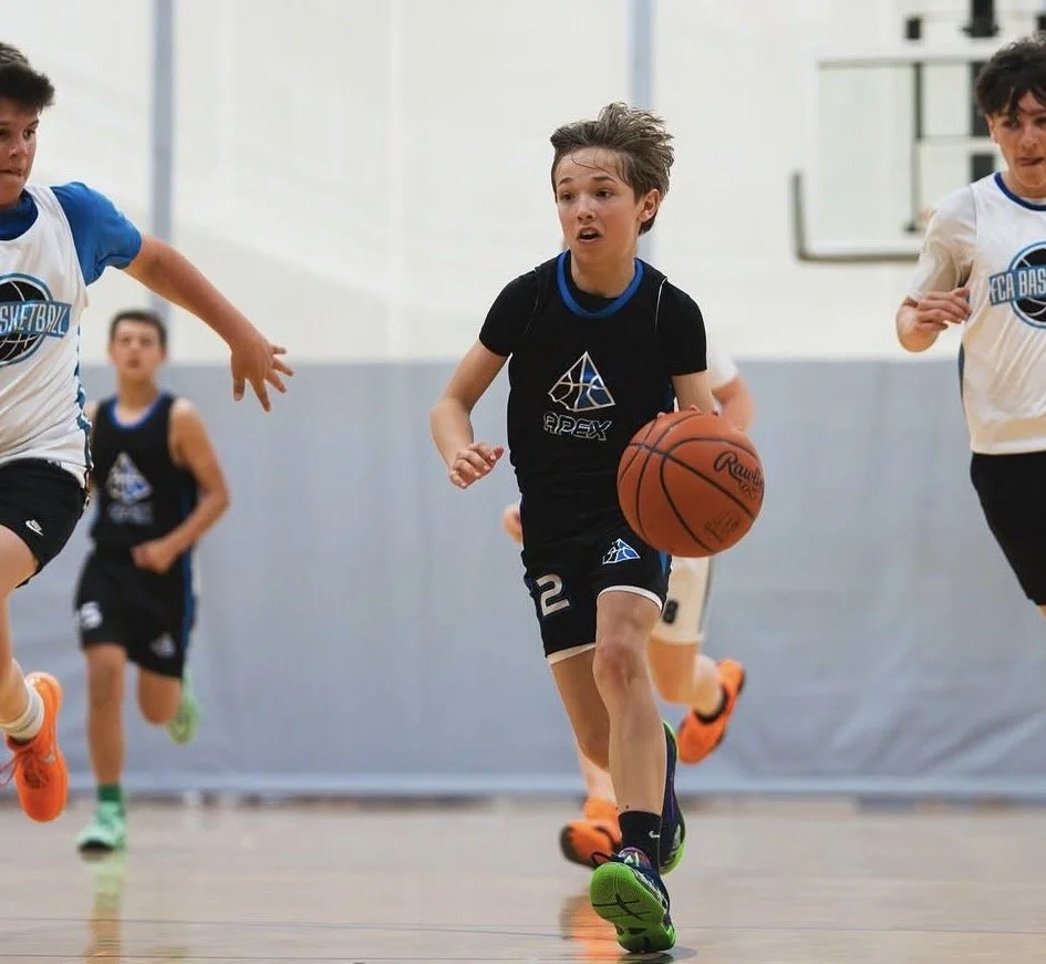 Young boys playing basketball on an indoor court, with one boy in black running with the ball while others are chasing him.