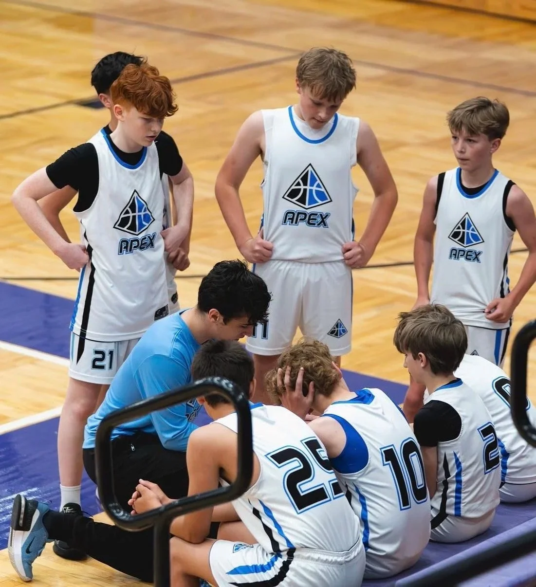 A boys' basketball team wearing white jerseys with the logo 'APEX' listens to their coach, who is crouched down and explaining something, as some players sit on the floor and others stand around them.