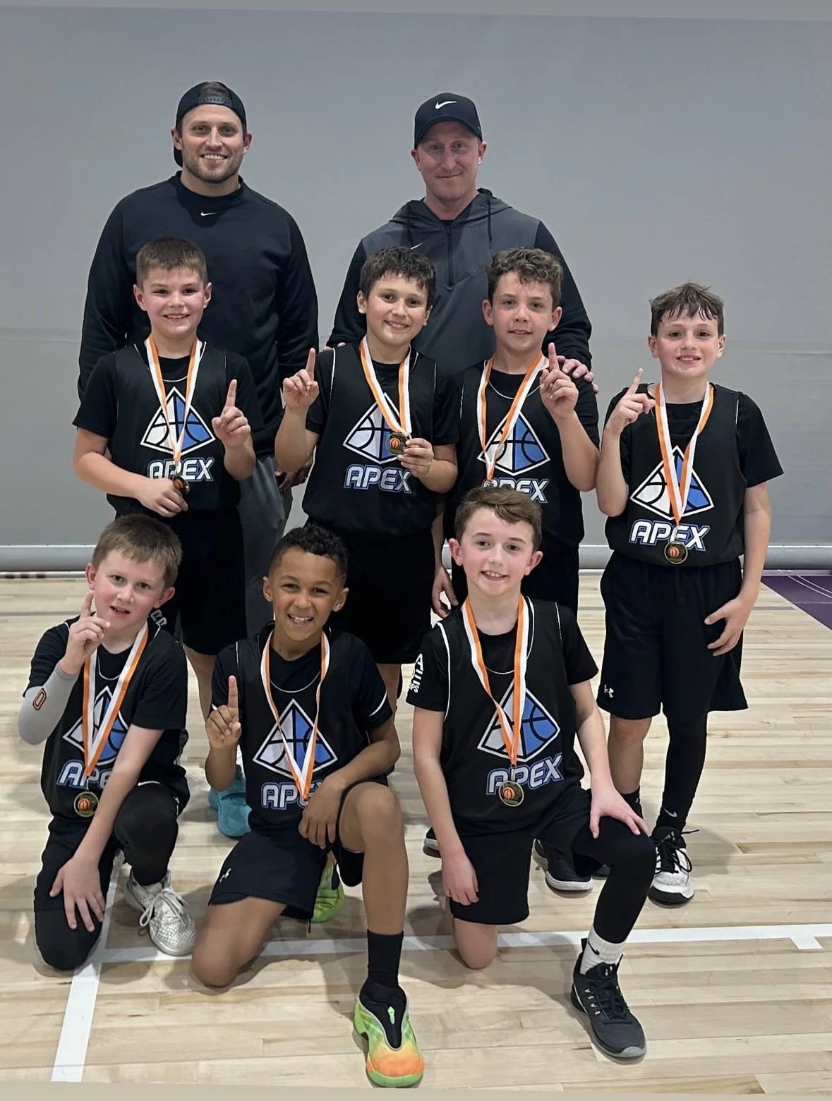 A youth basketball team of young boys, coached by two adult men, poses for a photo on an indoor basketball court. The boys are wearing black jerseys with a blue and white logo that says 'APEX' and are all wearing medals around their necks, indicating a recent victory or tournament win. The team members are smiling and holding up one finger, signifying they are number one.