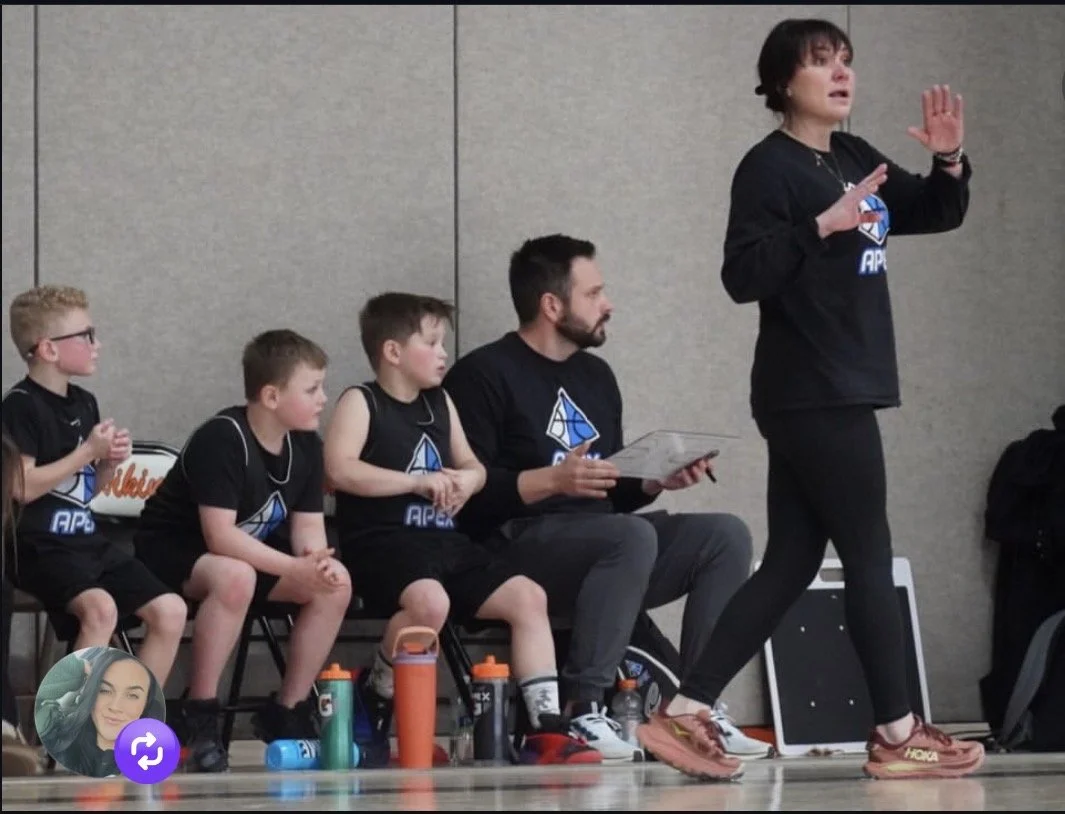A woman coaches a youth basketball team during a timeout, with four young players and a man seated on the sideline in matching black team shirts with a blue and white logo.