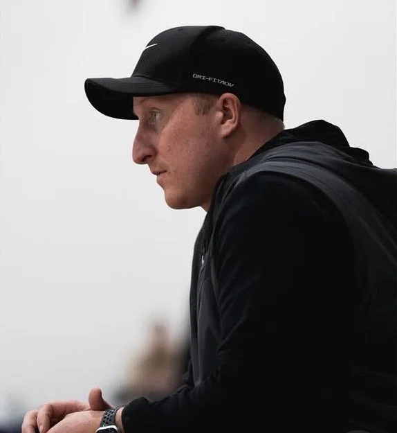 Side view of a man wearing a black Nike cap and black jacket, looking downward, with a blurred background.