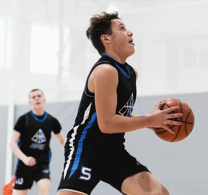 Two young boys playing basketball indoors. The boy in the foreground is holding a basketball and appears to be preparing to shoot or pass, wearing a black sleeveless basketball jersey with blue and white accents. The boy in the background is also dressed in a similar jersey, standing in the gym.