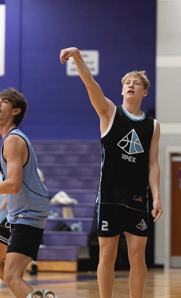 A young male basketball player in a black and blue jersey with the number 2, practicing a shot in a gymnasium, with another player in a light blue jersey nearby.