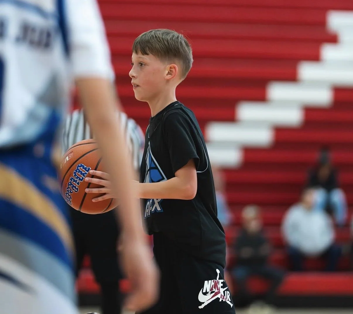 A young boy with short brown hair in a black T-shirt with a basketball graphic is holding a basketball at an indoor basketball court. Red and white bleachers are in the background with a few out-of-focus spectators.