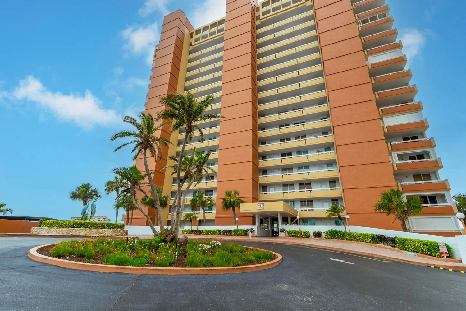 Exterior photograph of St. Petersburg condo in Pinellas County Florida. Cluster of palm trees in the foreground of the parking lot, and tall orange and yellow condo in the background.