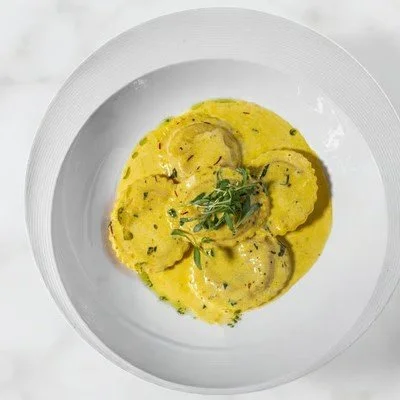 Plate of yellow ravioli with green herbs and black pepper in a creamy sauce.