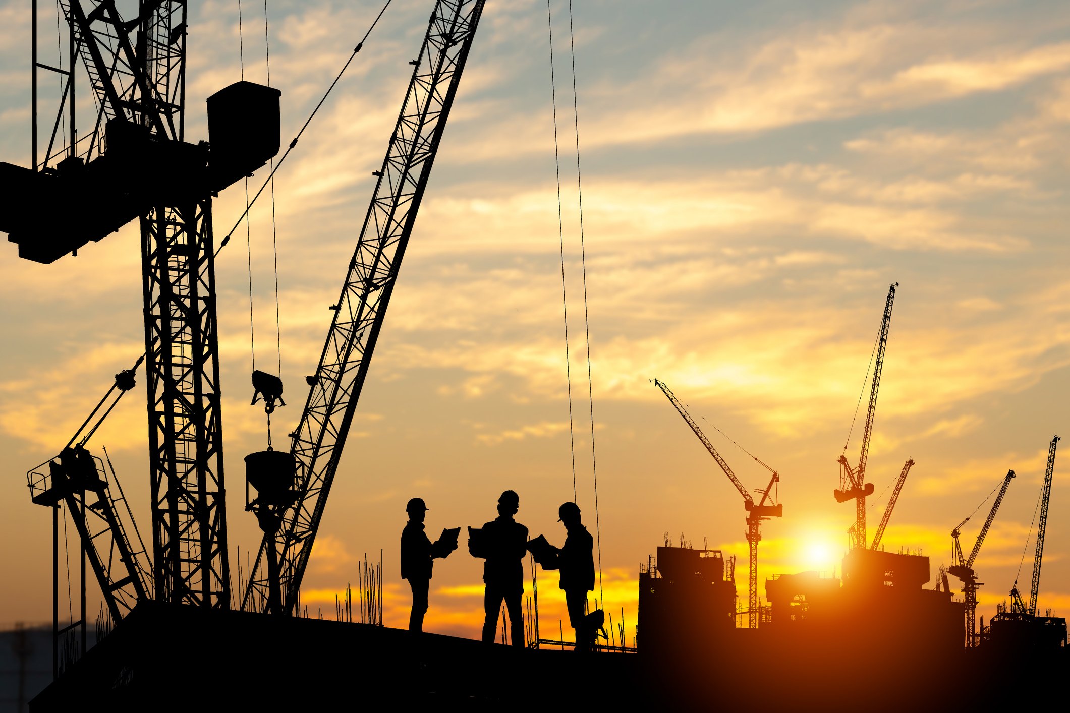 Silhouettes of construction workers and cranes at a construction site during sunset or sunrise.