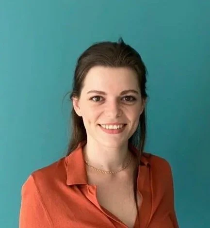 Une femme souriante avec des cheveux bruns, portant une chemise orange, contre un fond bleu.
