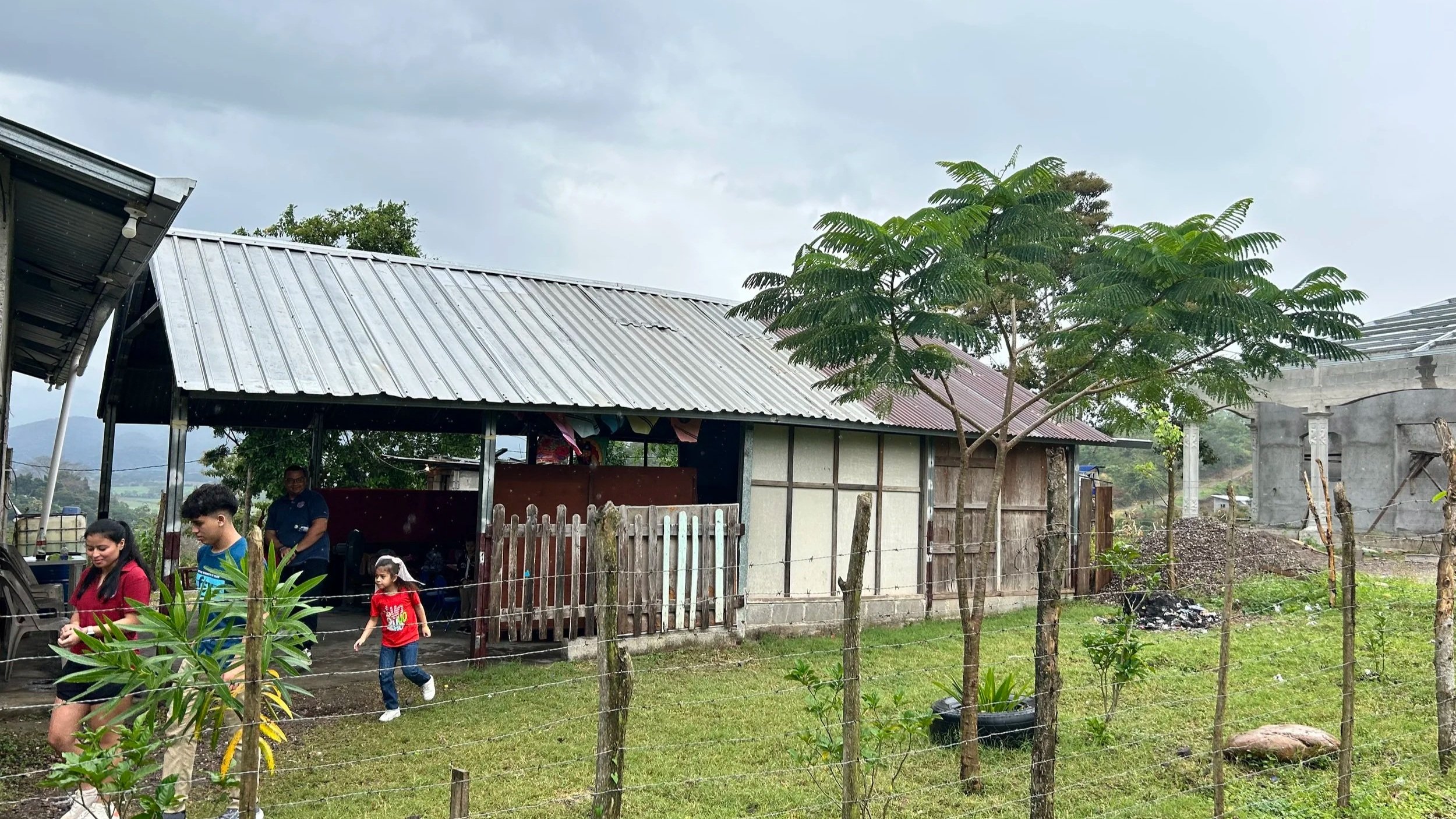 A rural scene with a small house having a metal roof, a young tree and a group of people, including children, outside near a fenced yard on a cloudy day.