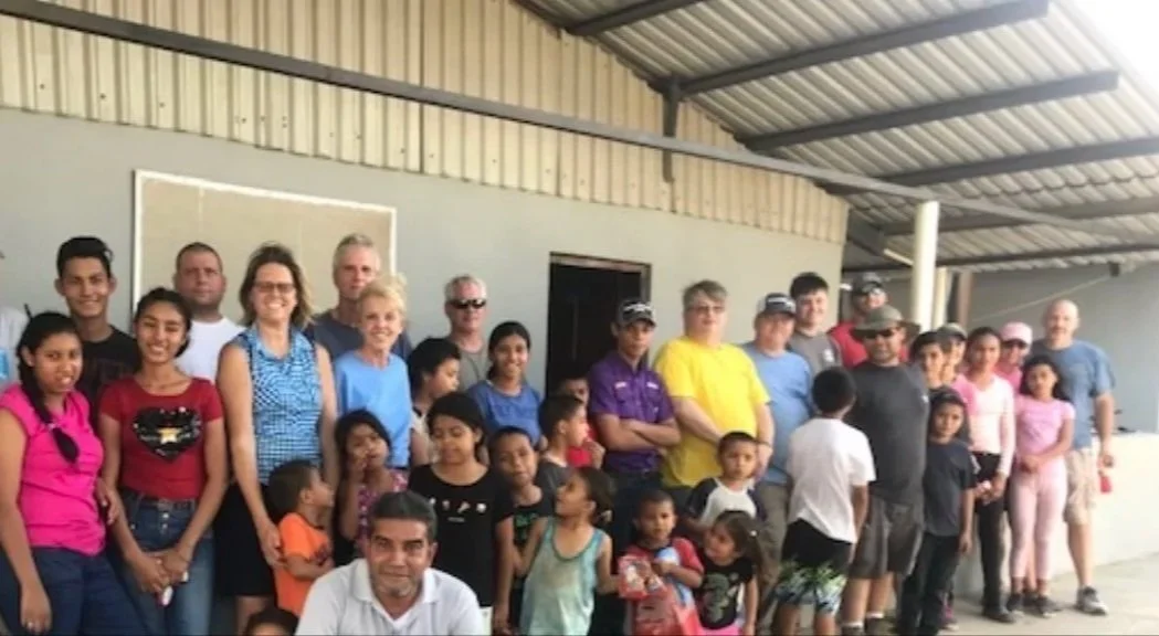 Group of adults and children gathered outside under a metal roof, posing for a photo.
