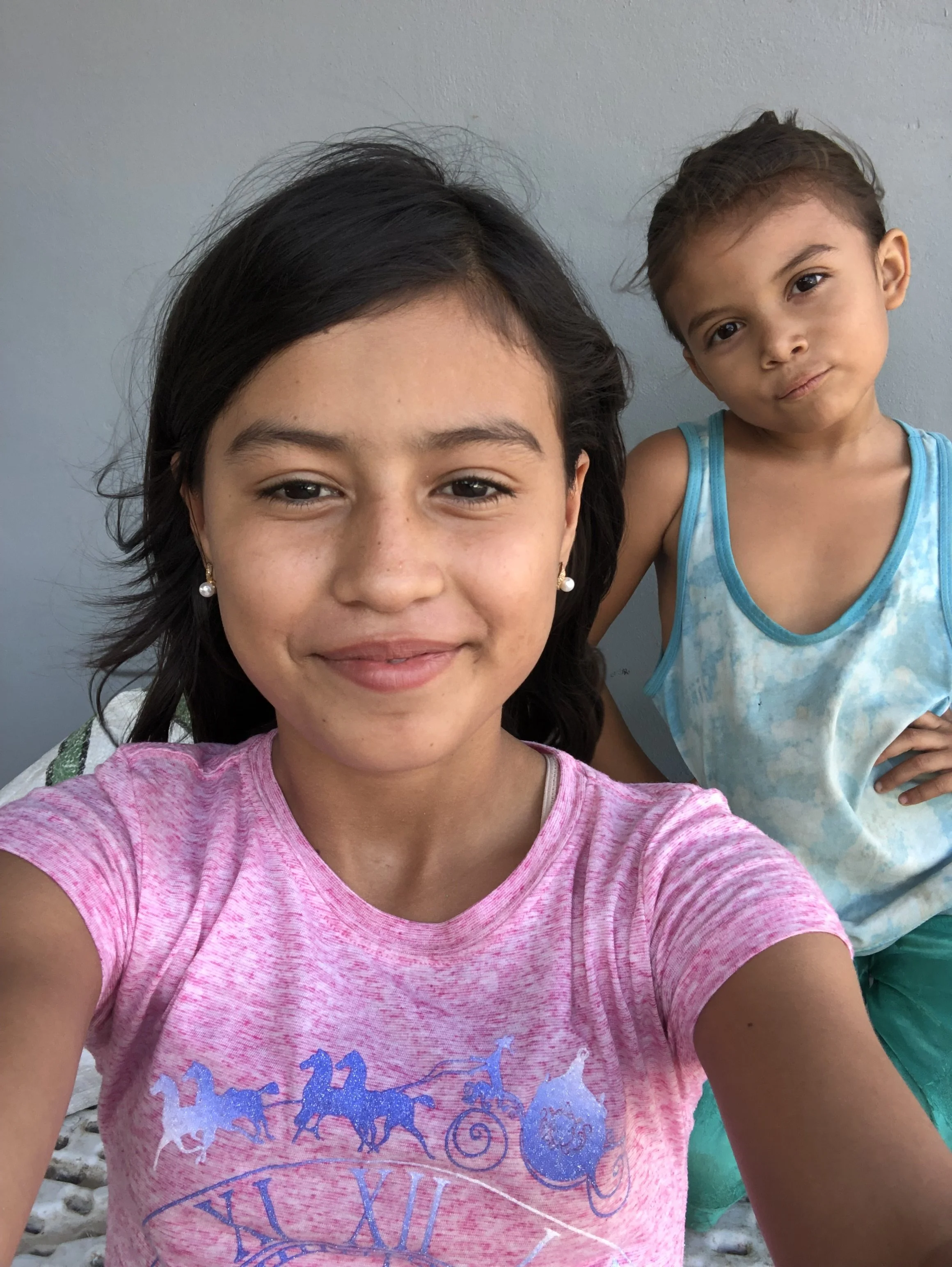 Close-up of two young girls taking a selfie against a plain wall. The girl in the foreground has dark hair, tan skin, and is wearing a pink shirt with a graphic design. The girl in the background has lighter skin, brown hair, and is wearing a blue an
