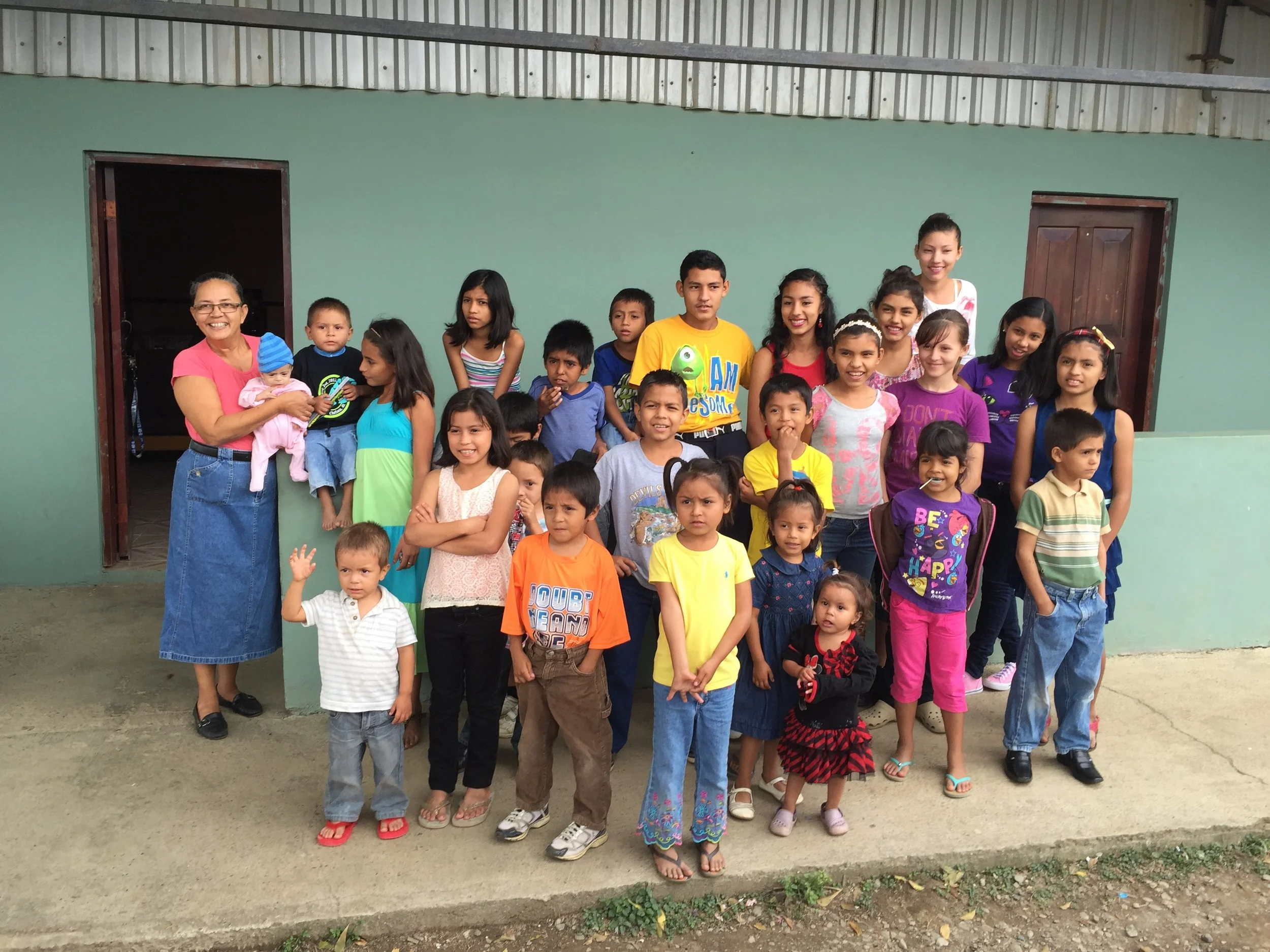 A group of children and a woman, possibly a teacher or caregiver, standing and smiling in front of a green wall with two open doorways. The children are of various ages, wearing colorful clothes, and appear to be outdoors or in a semi-open space.