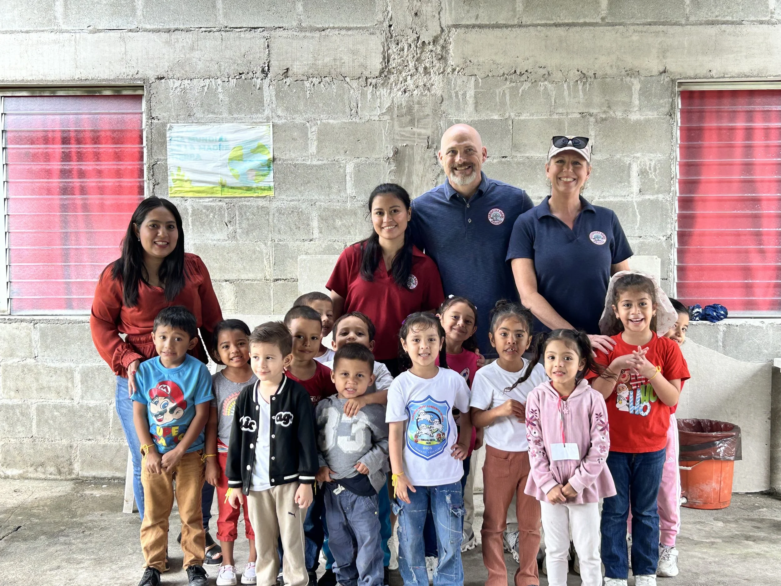 Group of young children and three adults, including two women and one man, standing together inside a building with concrete walls and windows with red shades. The children are smiling, and the adults are smiling at the camera.