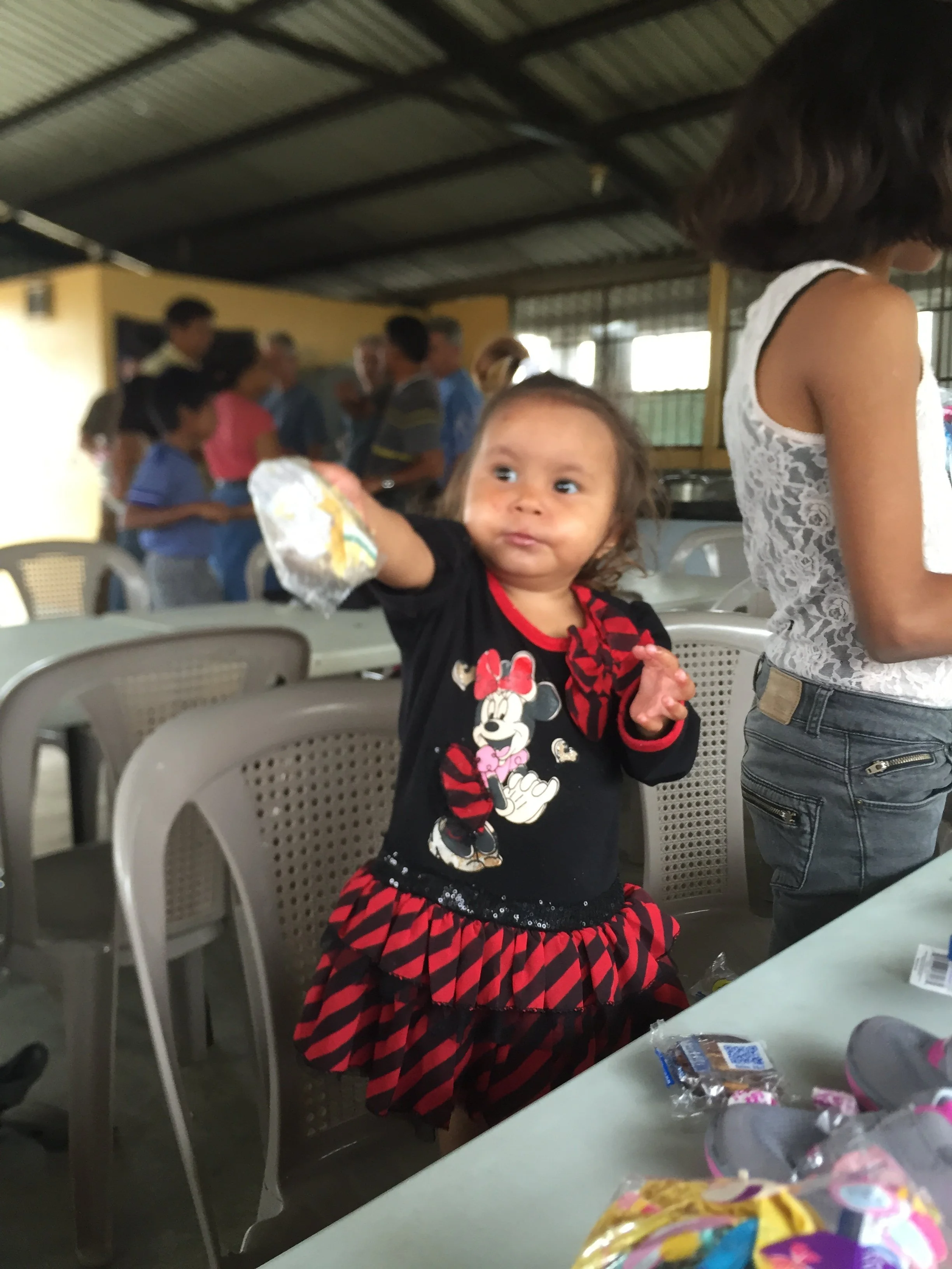 A young girl with long brown hair sitting at a table, wearing a black and red Minnie Mouse dress, looking to the side. Behind her, a woman in a white lace top and jeans stands, with her back partially visible. In the background, a group of people are