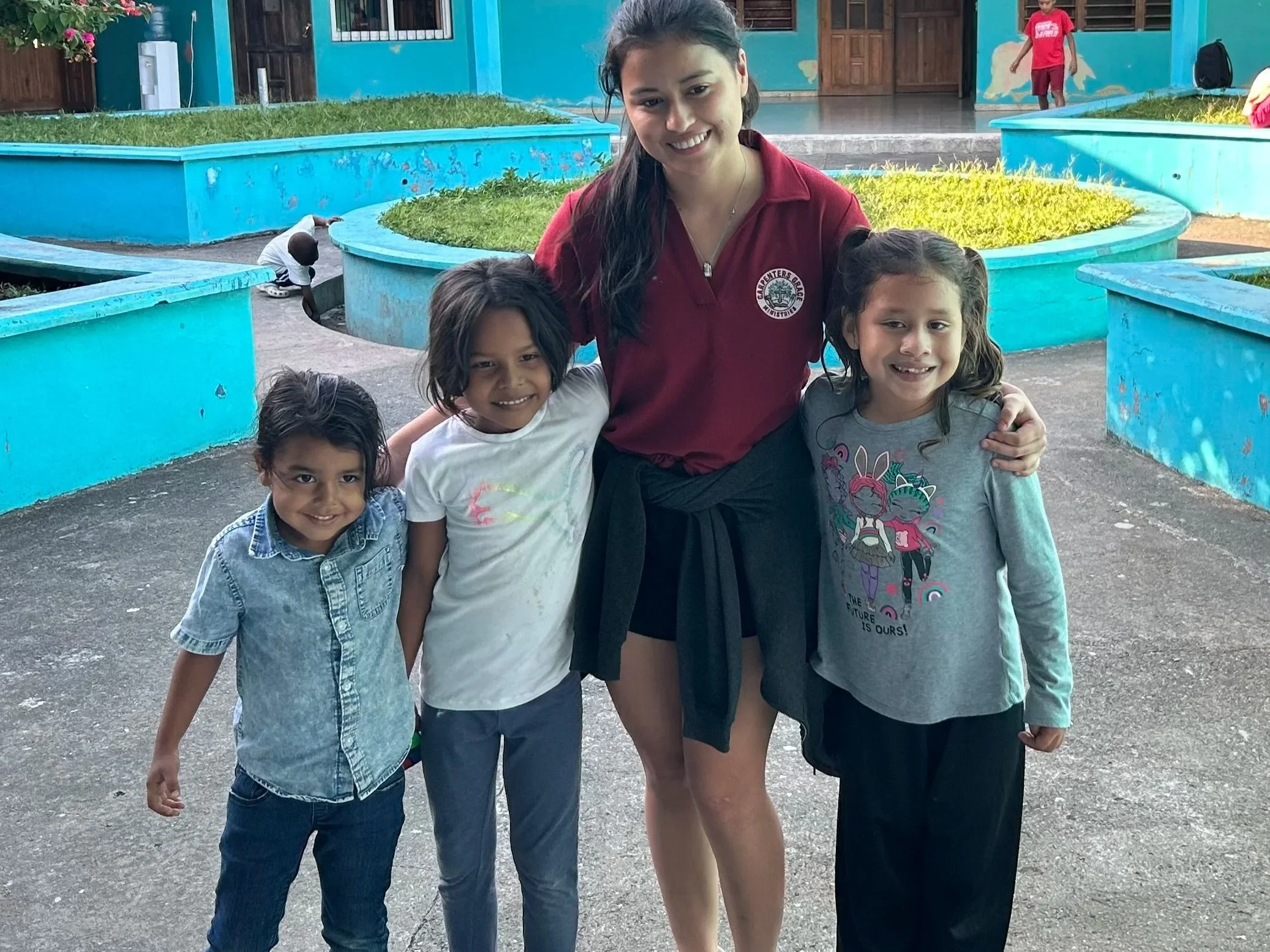 A woman in a maroon shirt with a logo, smiling, standing outdoors with four young girls, all smiling, in front of painted blue structures and greenery.