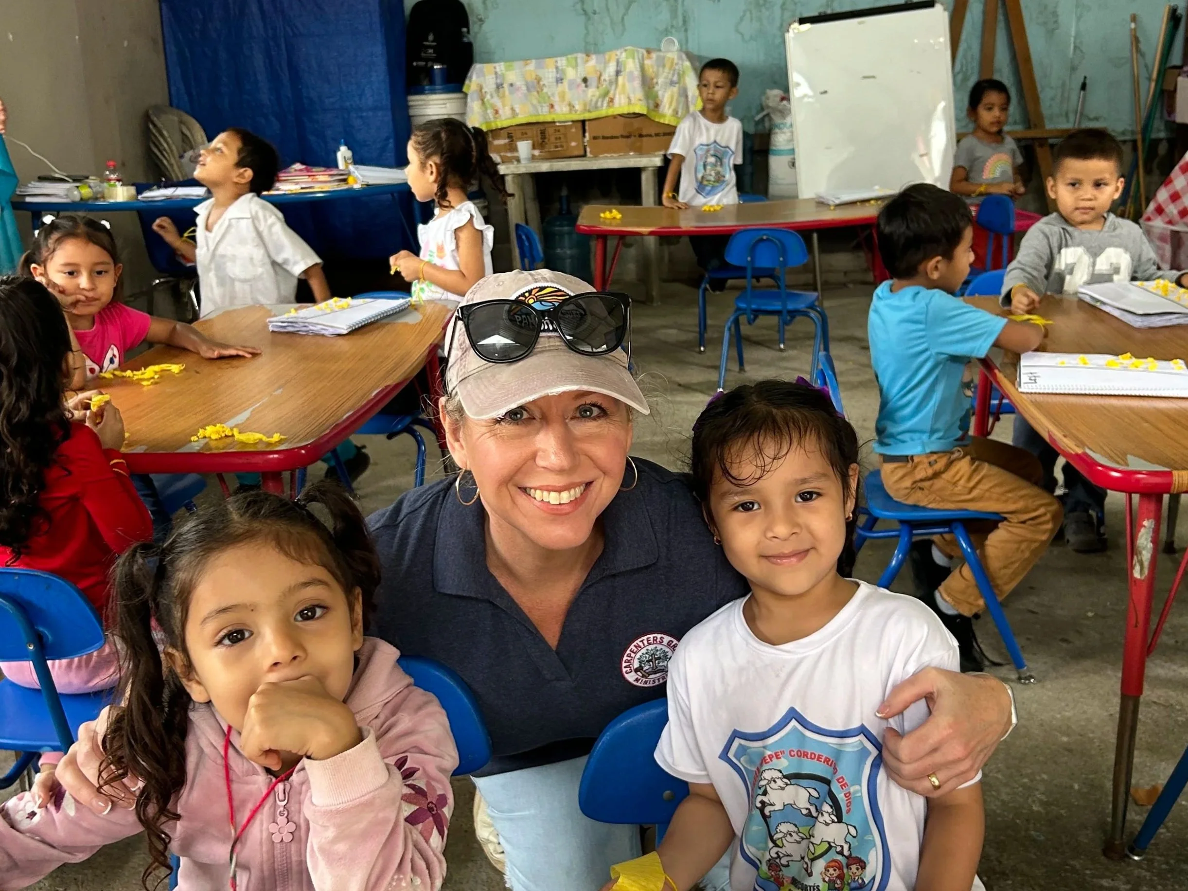 A group of young children and an adult woman smiling and posing for the camera in a classroom with various children in the background. The children are seated at tables with some wearing casual clothes and some with school shirts. The woman is wearing a baseball cap and sunglasses on top of her head.