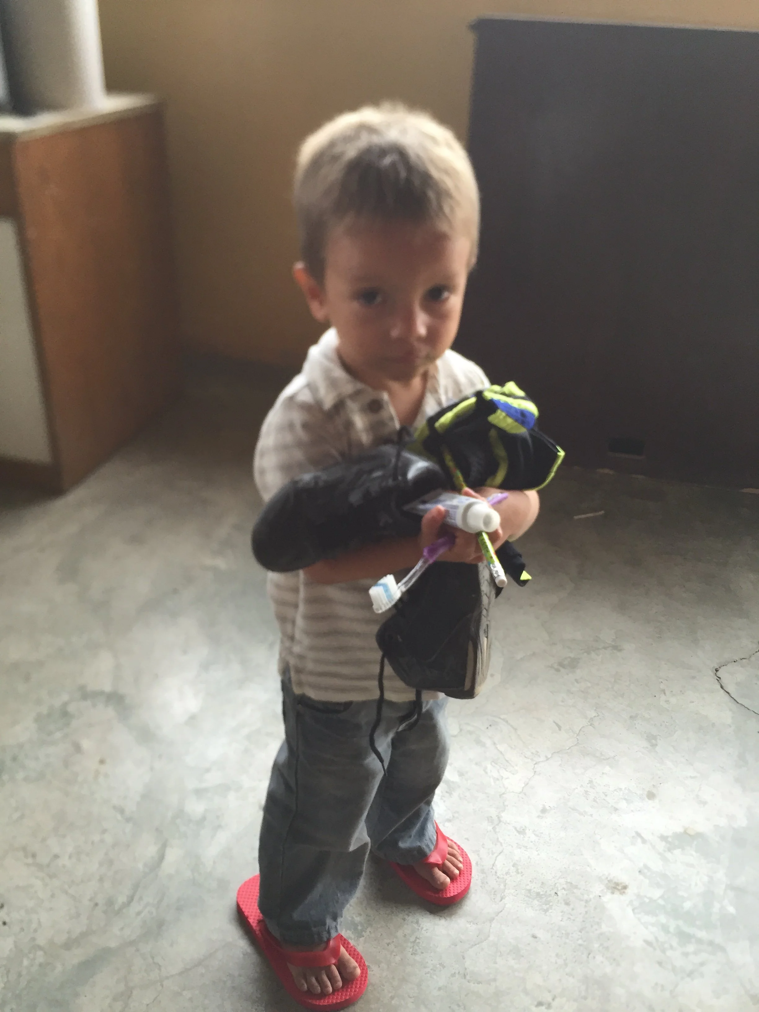 A young boy with light skin and short blond hair is standing on a kitchen floor, wearing red flip-flops, gray pants, a plaid shirt, and holding some items including a water bottle and a toy. He is looking at the camera with a neutral expression.