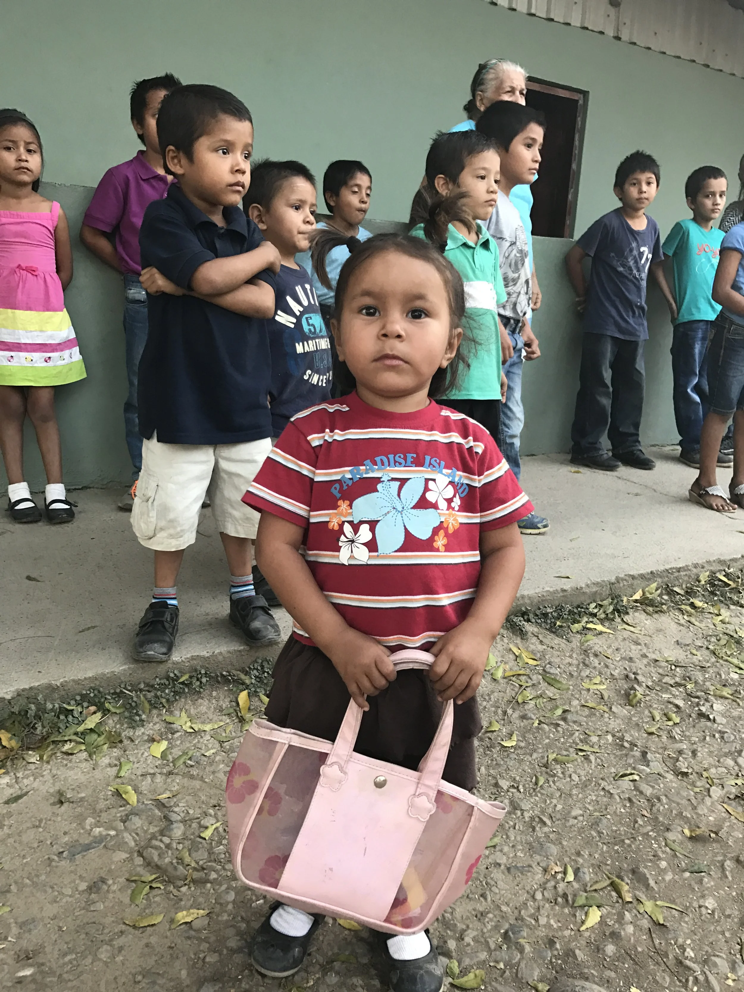 A young girl standing in front of a group of children and an elderly woman. The girl is holding a pink bag with a floral design and is wearing a red shirt with colorful flowers. The children are lined up against a green wall, wearing casual clothes, 