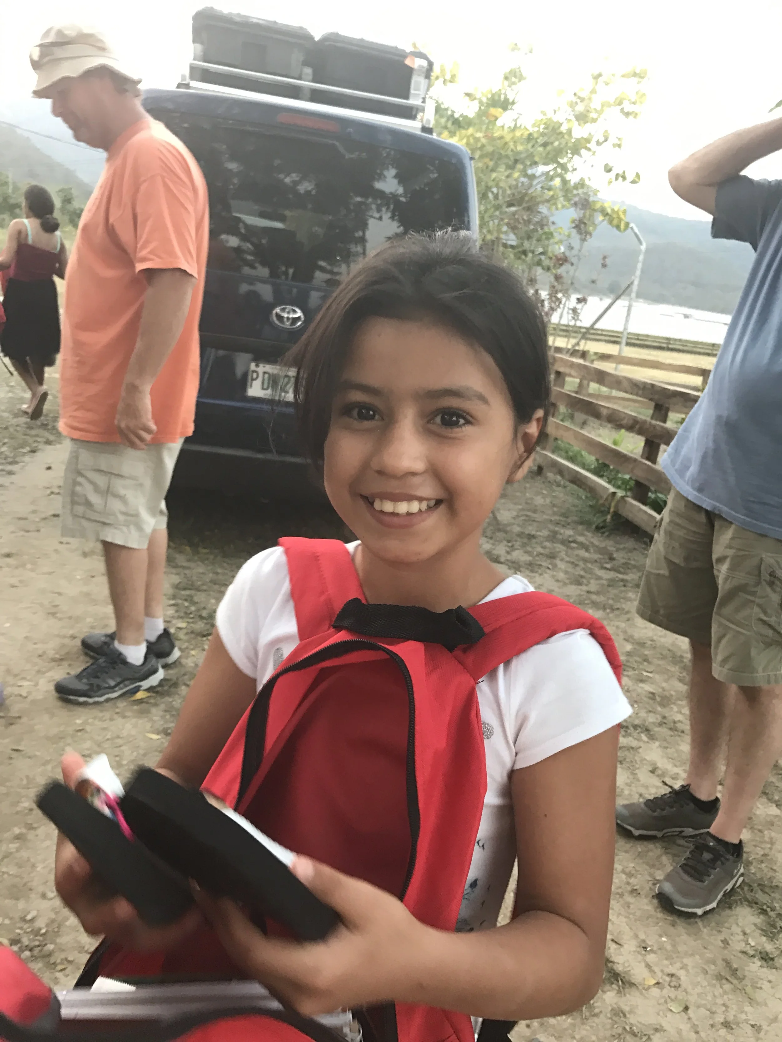 A young girl with dark hair smiling at the camera, wearing a white shirt with a red backpack. Several people are outdoors near a black Toyota vehicle, trees, and a wooden fence in the background, under a cloudy sky.