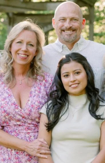 A family of three posing outdoors, with trees and a wooden fence in the background. The woman on the left has blonde hair and is wearing a pink floral dress. The man on the right has a bald head, beard, and is wearing a light-colored collared shirt. The young woman in front has long dark hair and is wearing a light-colored top.