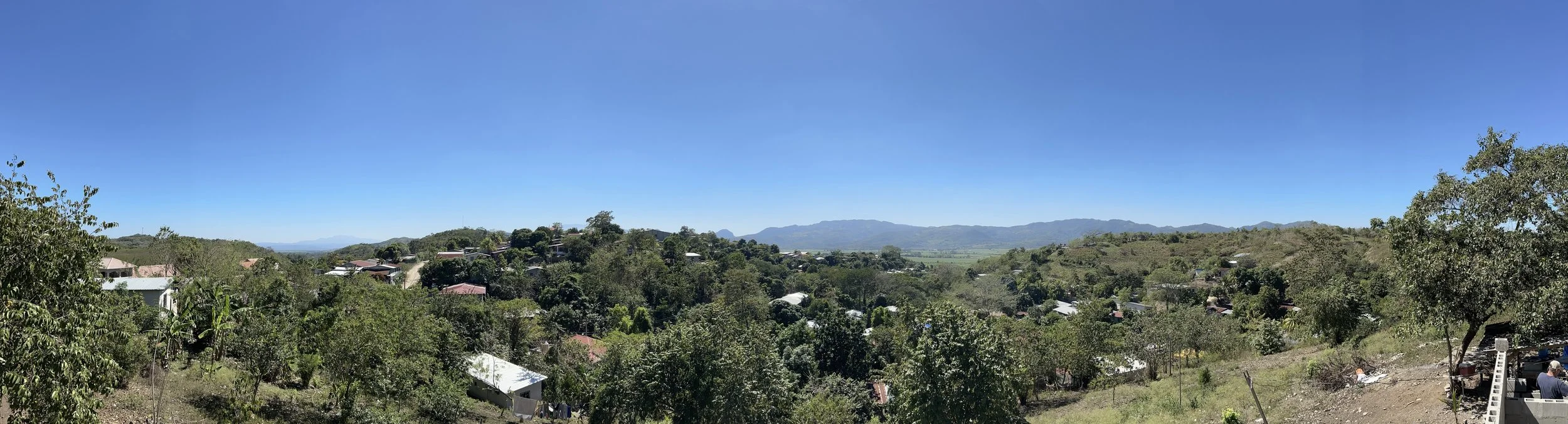 Panoramic view of a rural landscape with houses, trees, and distant mountains under a clear blue sky.