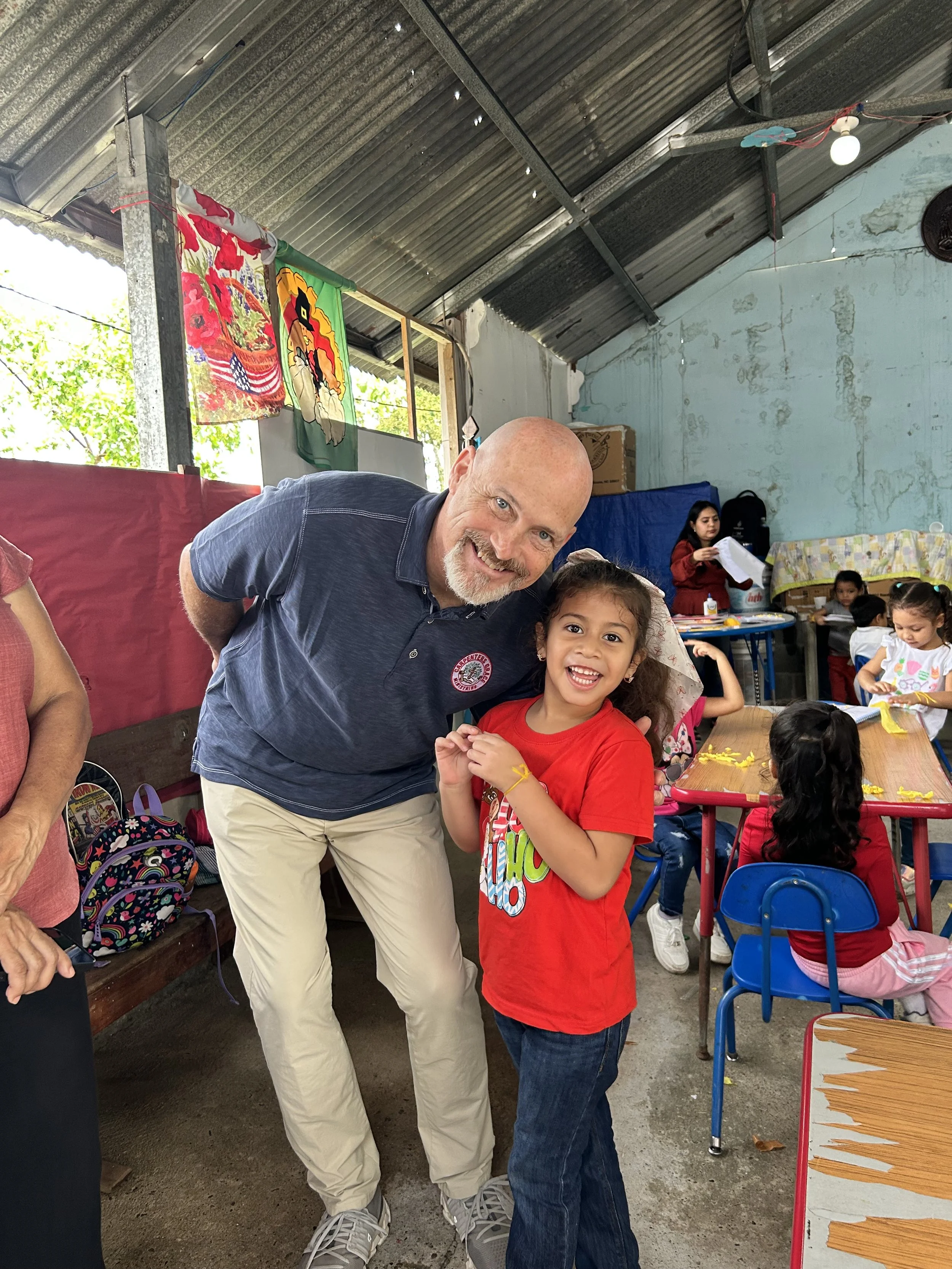 A man with a bald head and a beard smiling and leaning towards a young girl with curly hair, also smiling, in a classroom with other children and teachers in the background.