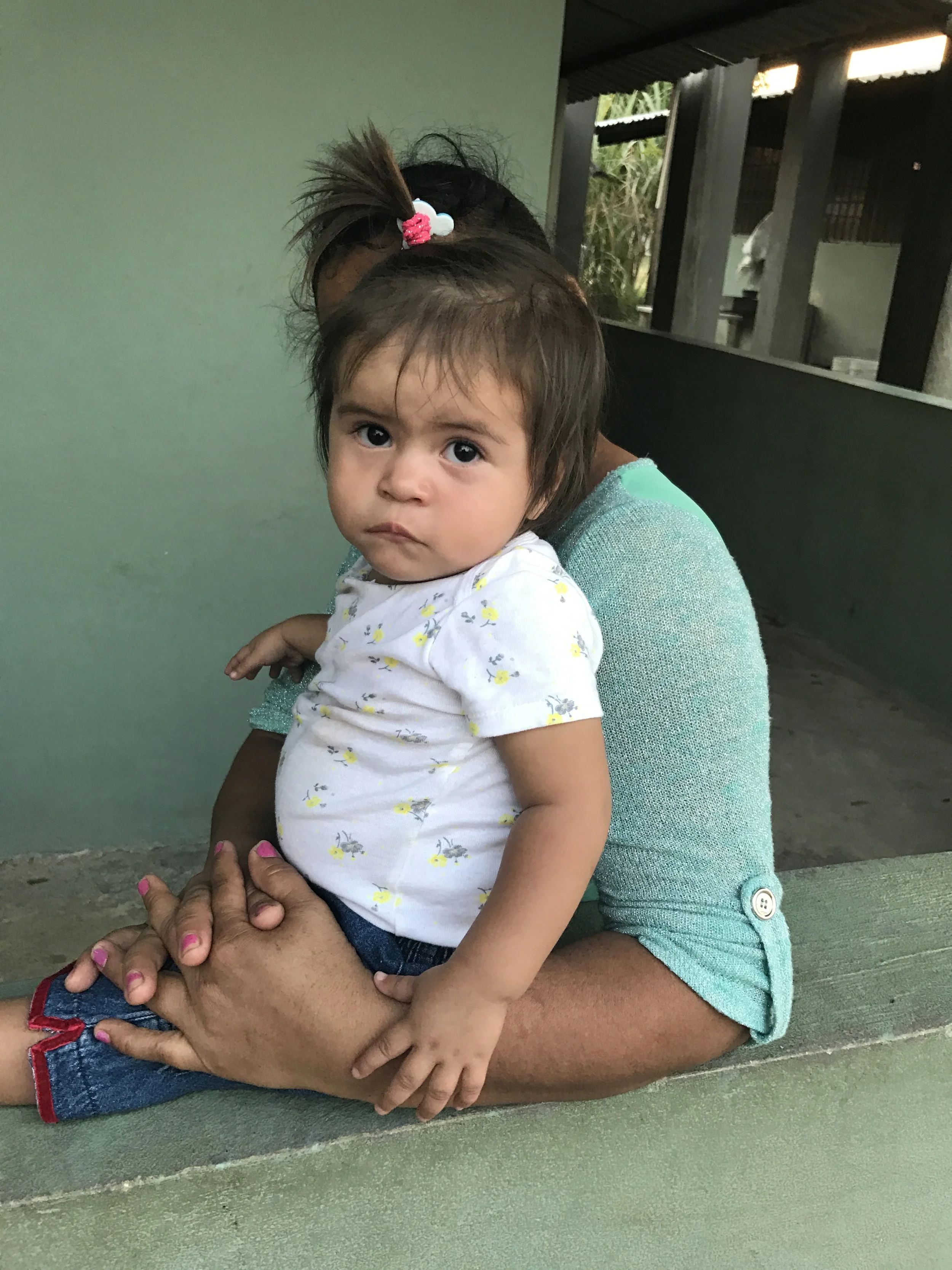 A young girl with brown hair tied with a pink hairband, sitting on an adult's lap. The adult's arms are visible, hugging the girl, with pink nail polish on the nails. The girl is wearing a white shirt with yellow and gray patterns. They are sitting o