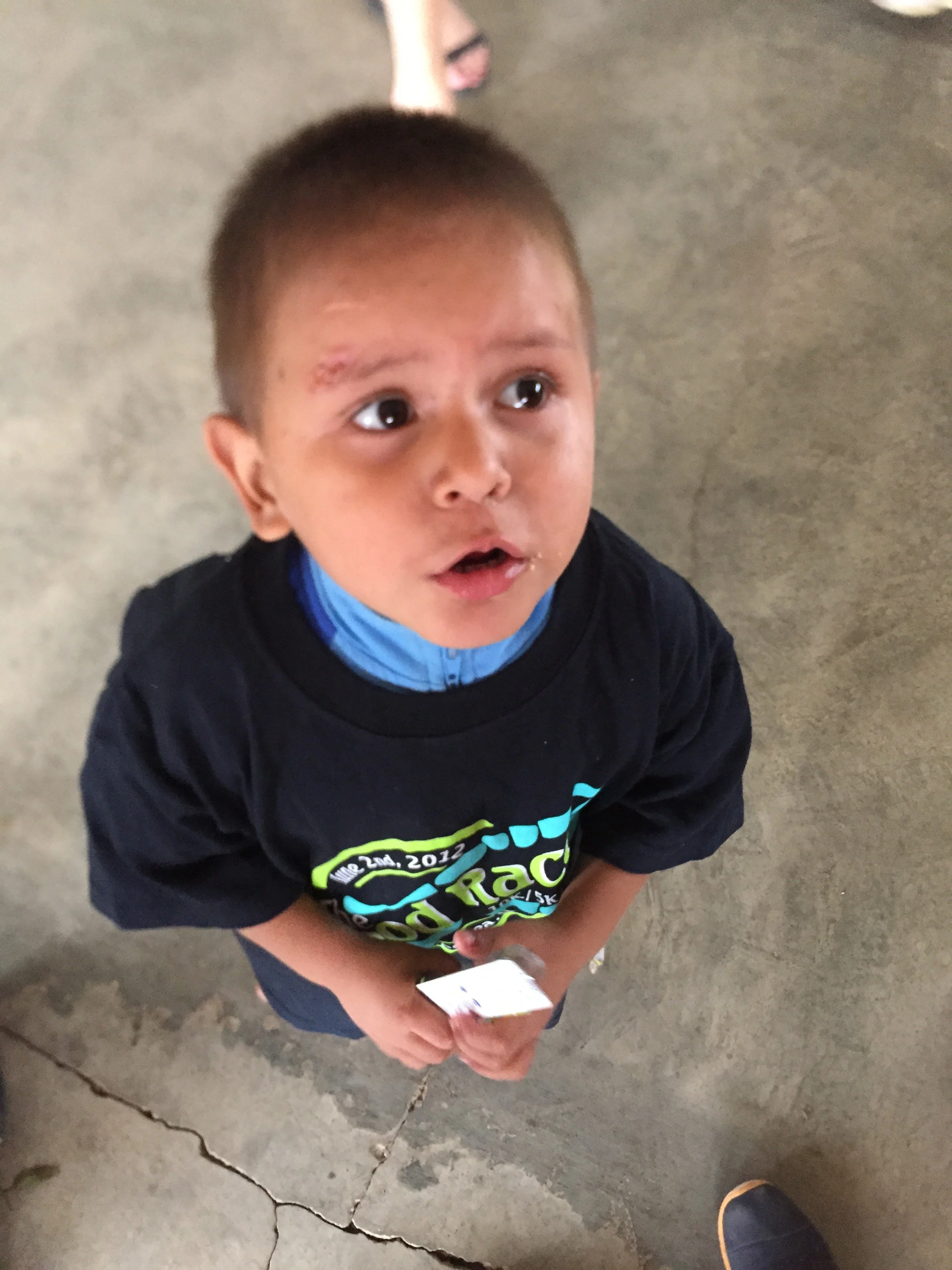 A young boy with short dark hair and a small scrape on his forehead, wearing a black t-shirt with colorful text, standing on a cracked concrete floor, holding a piece of paper and looking up, with adult feet visible in the background.