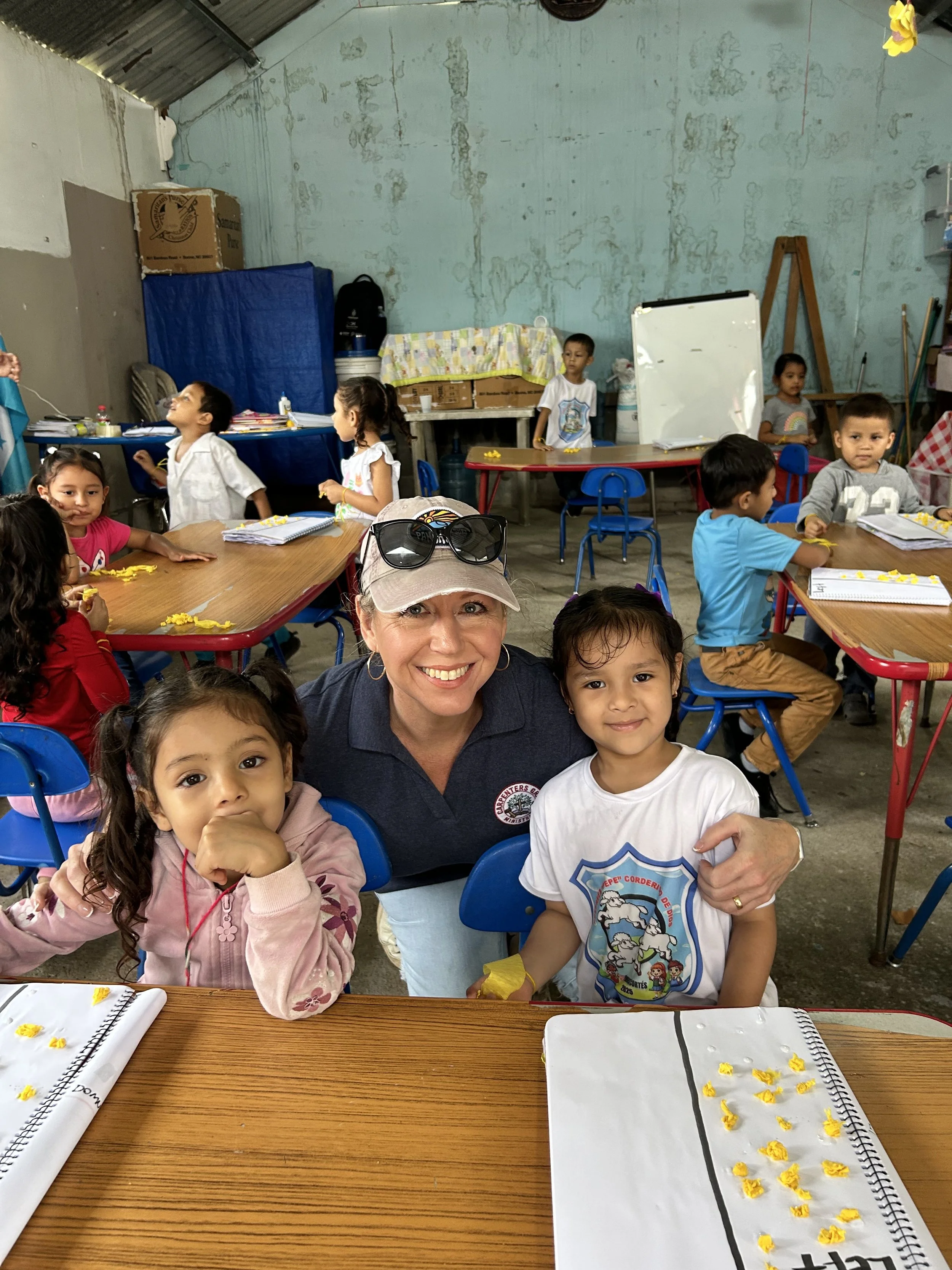 A group of children and a woman smiling and sitting at tables inside a room with a corrugated metal roof, blue and red chairs, some tables have notebooks and yellow paper decorations, a woman is taking a selfie