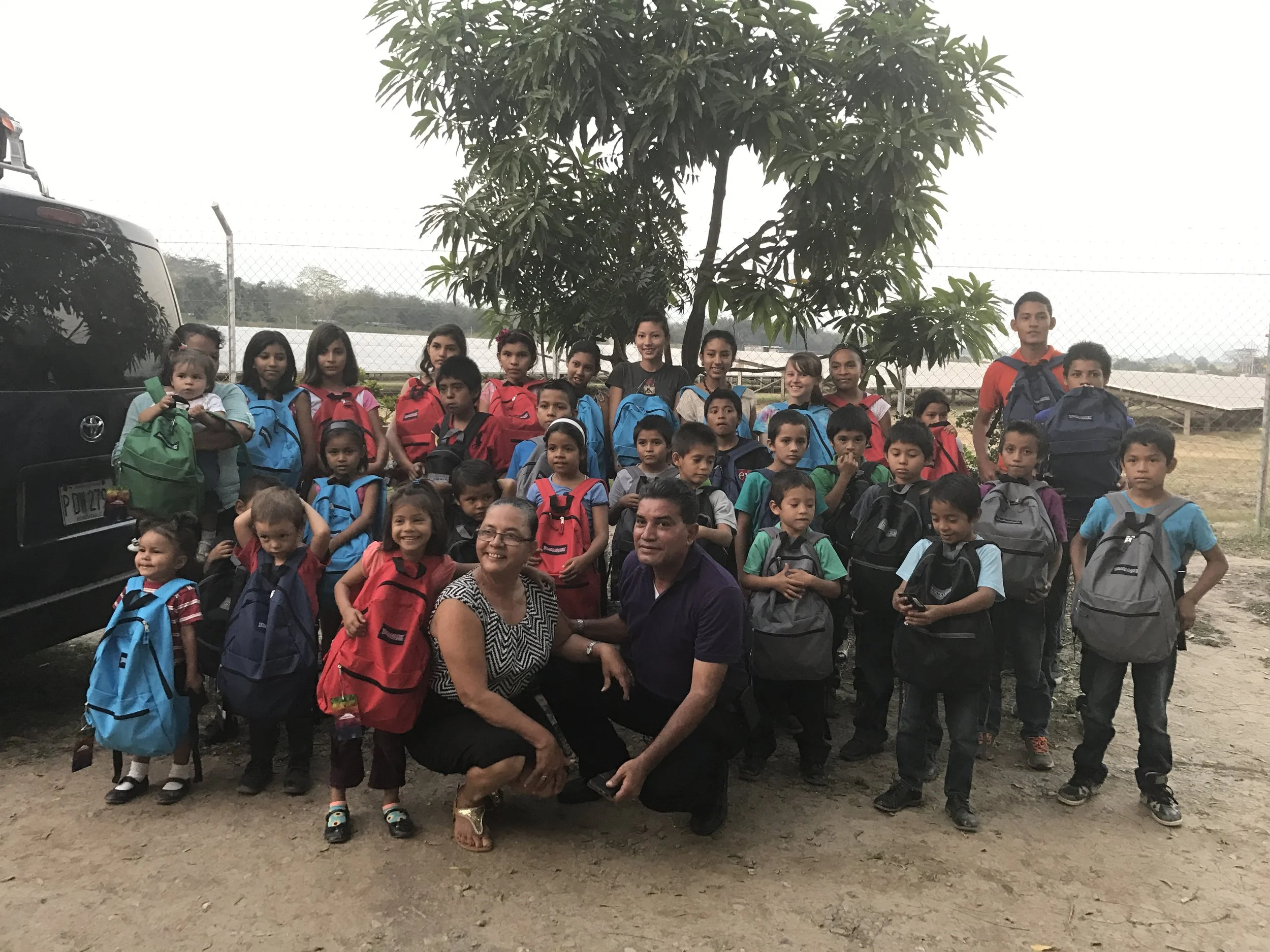 Group of children and adults outdoors near a tree, some children wearing backpacks, standing on dirt ground, with a car and fence in the background.