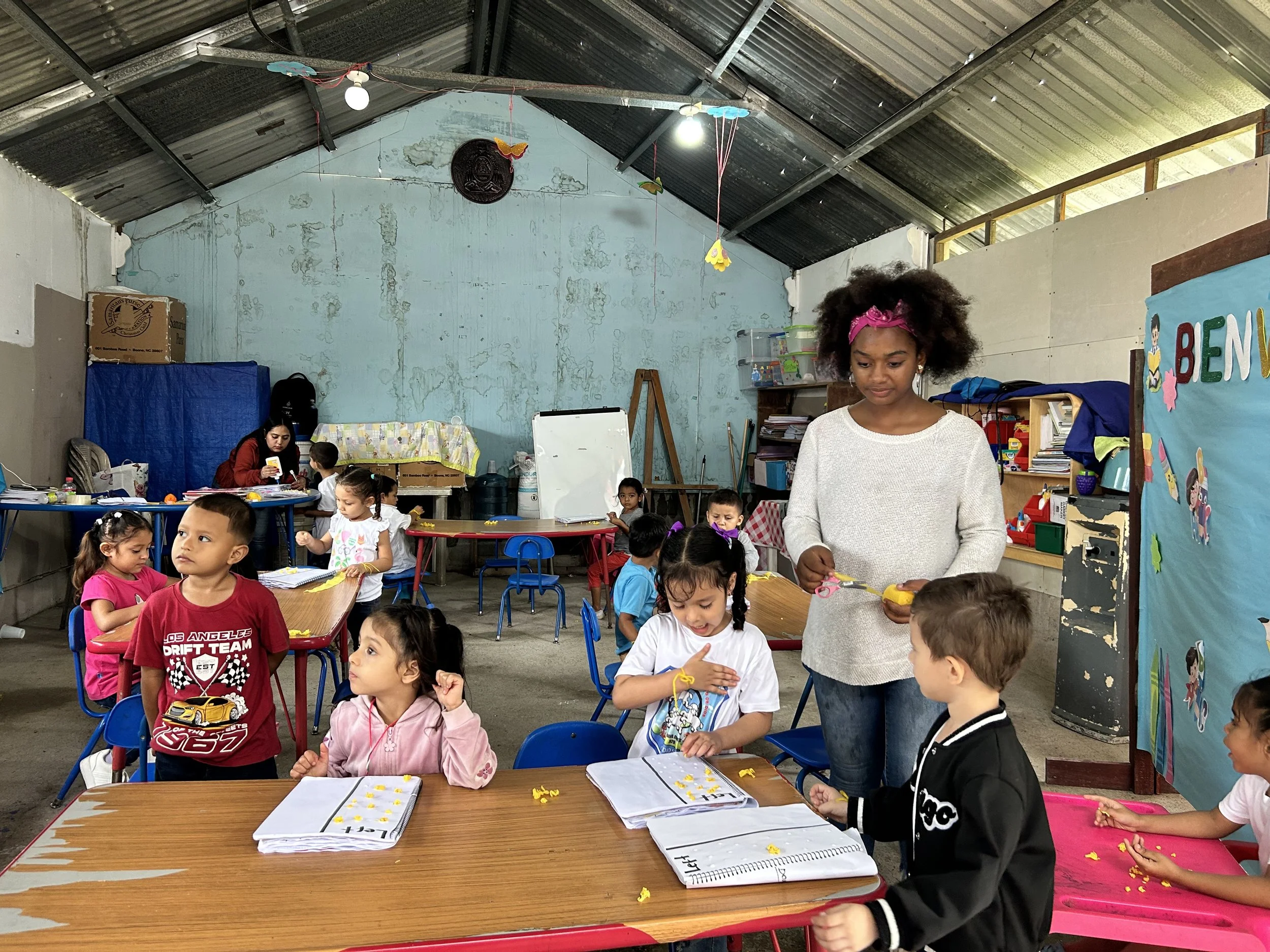 Children sitting at tables doing arts and crafts in a classroom with a woman supervising.
