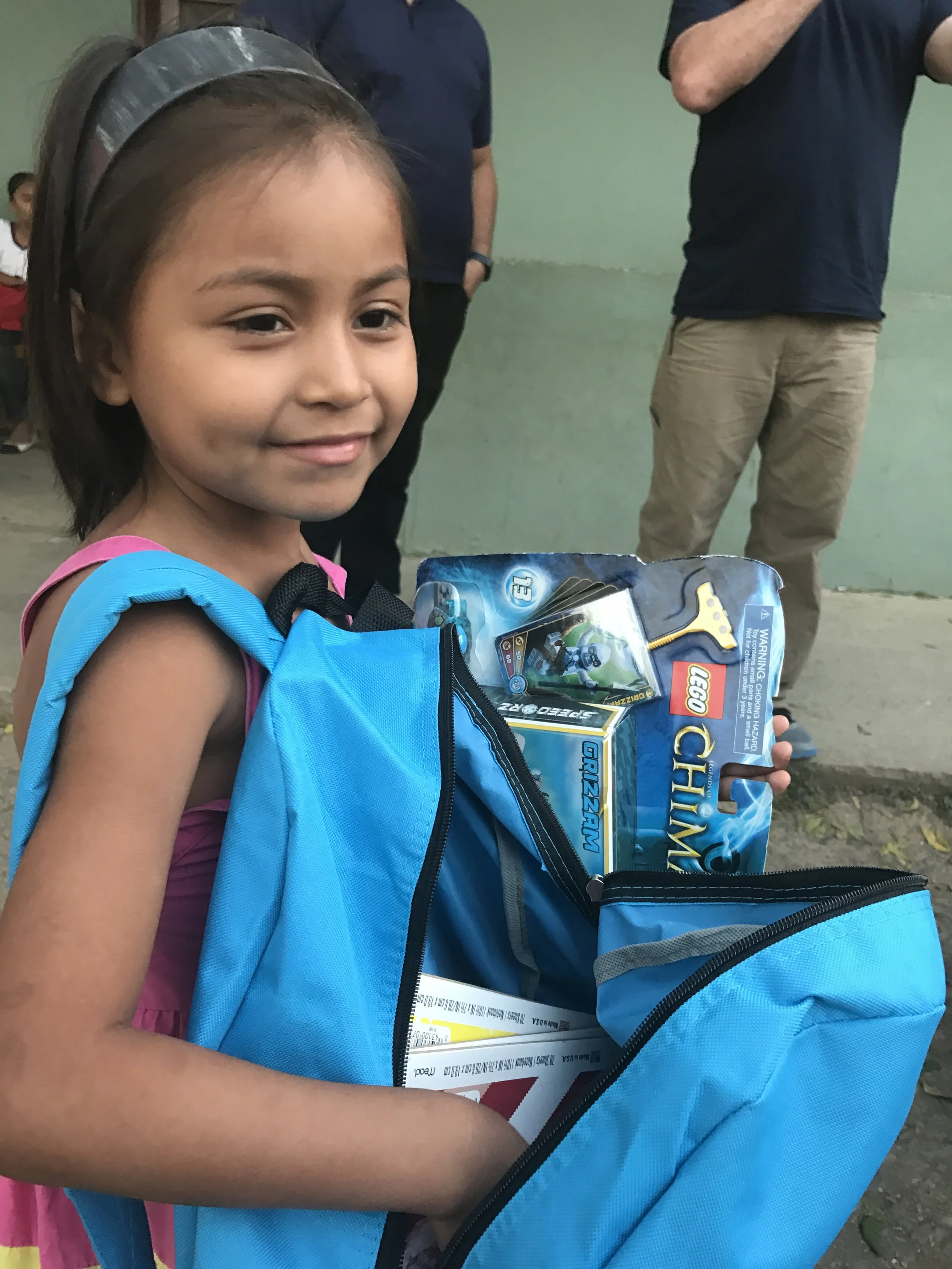 A young girl with a blue backpack and a black headband holding a LEGO construction toy set and some magazines, smiling, standing outdoors with a green wall and two adults in the background.