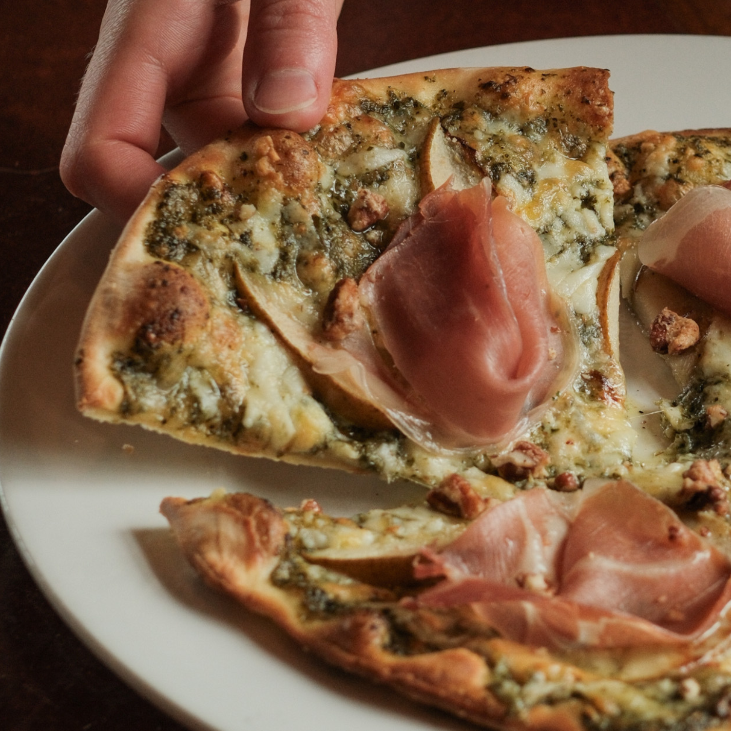 Close-up of a pizza with cheese, herbs, and slices of ham on a white plate, being held by a hand.