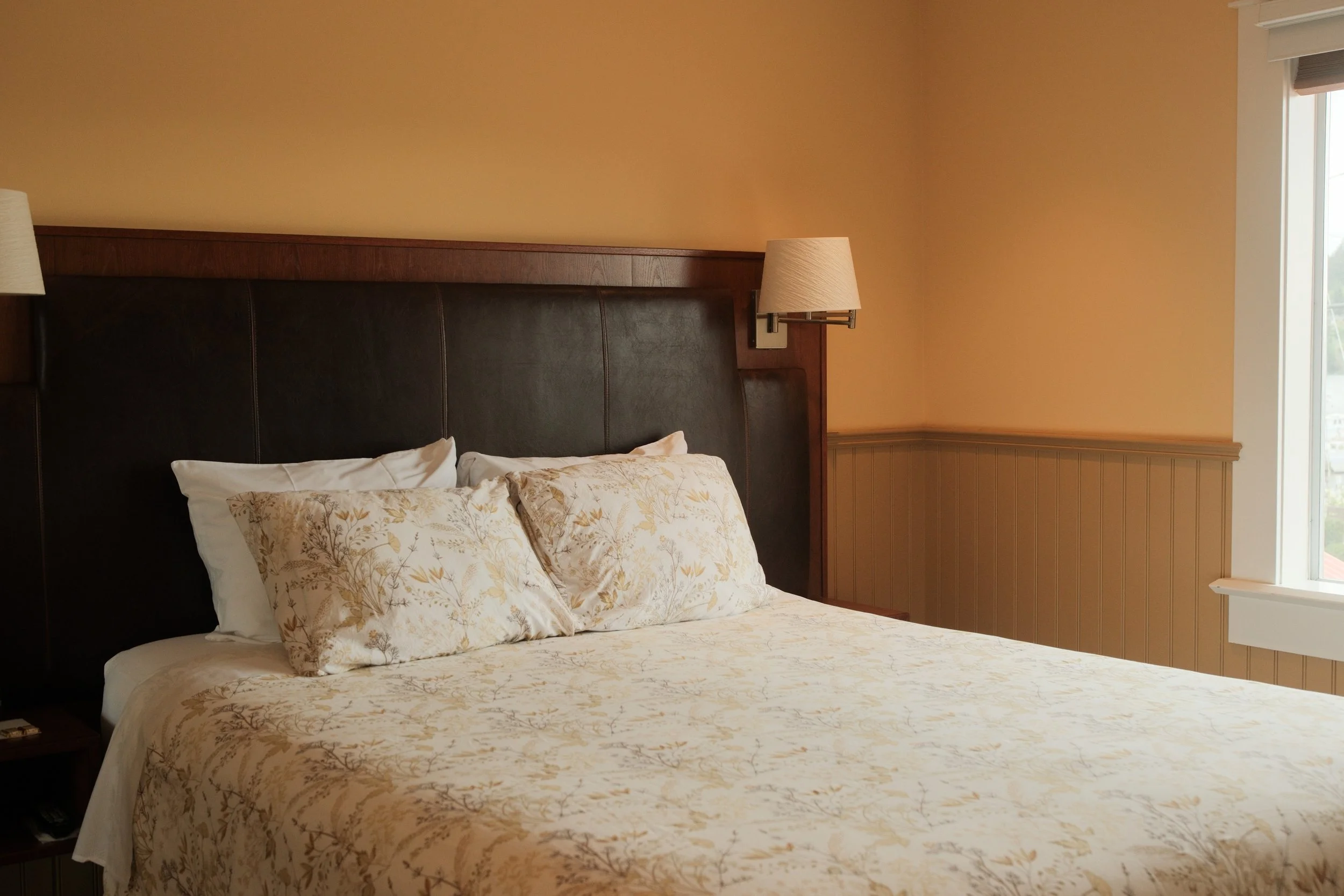 A neatly made bed with floral patterned bedding and pillows in a cozy bedroom with warm walls, a wall-mounted lamp, and a window with blinds letting in natural light.