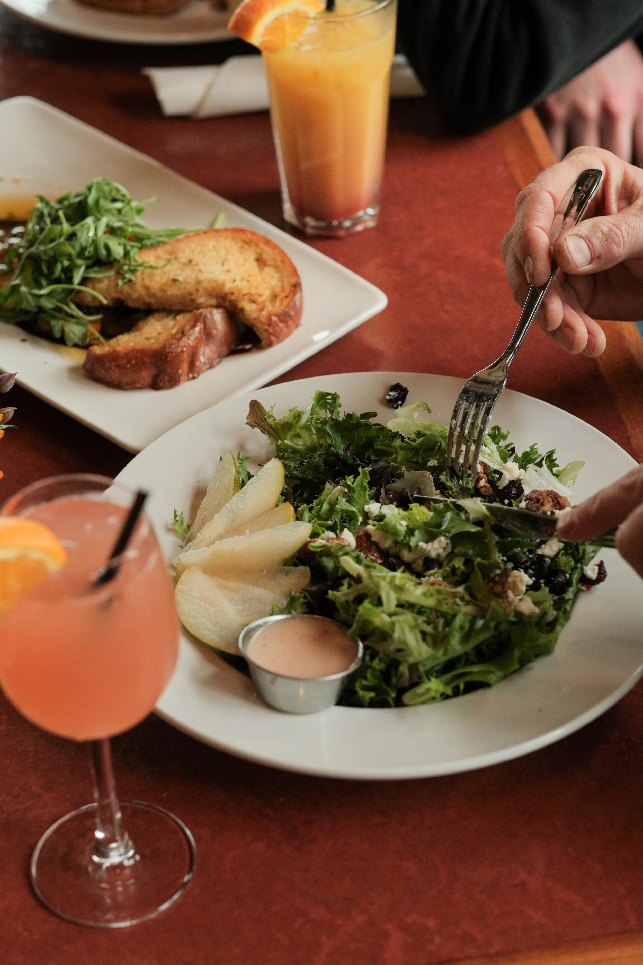 A glass of orange juice with a slice of orange on the rim, a plate of salad with greens, cheese, and dark berries, a dish of breaded chicken with greens, and a pink cocktail with a slice of orange.