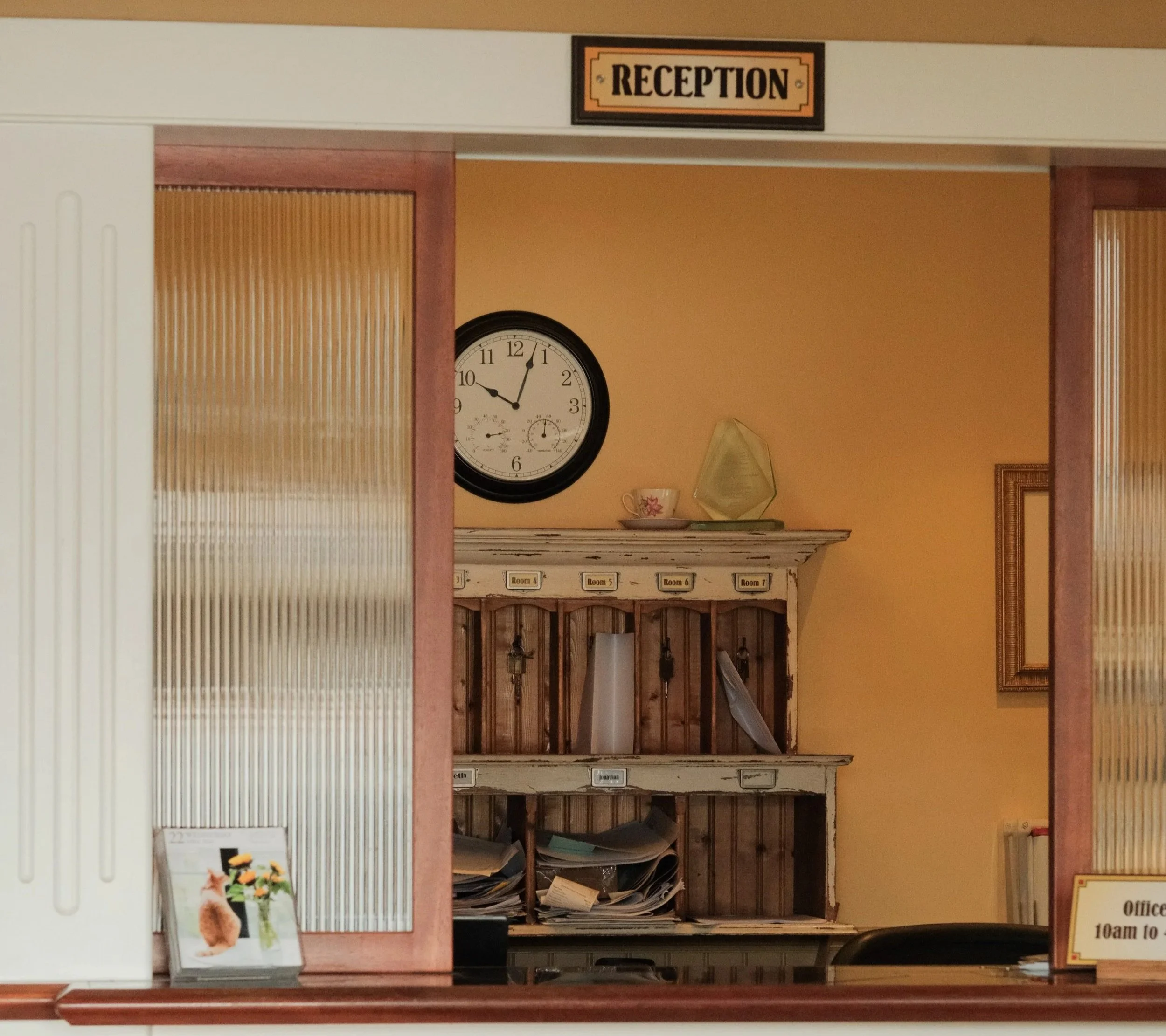 Reception area with wooden shelves filled with papers, a clock on the wall showing 10:05, and a sign indicating office hours from 10 am to 5 pm.