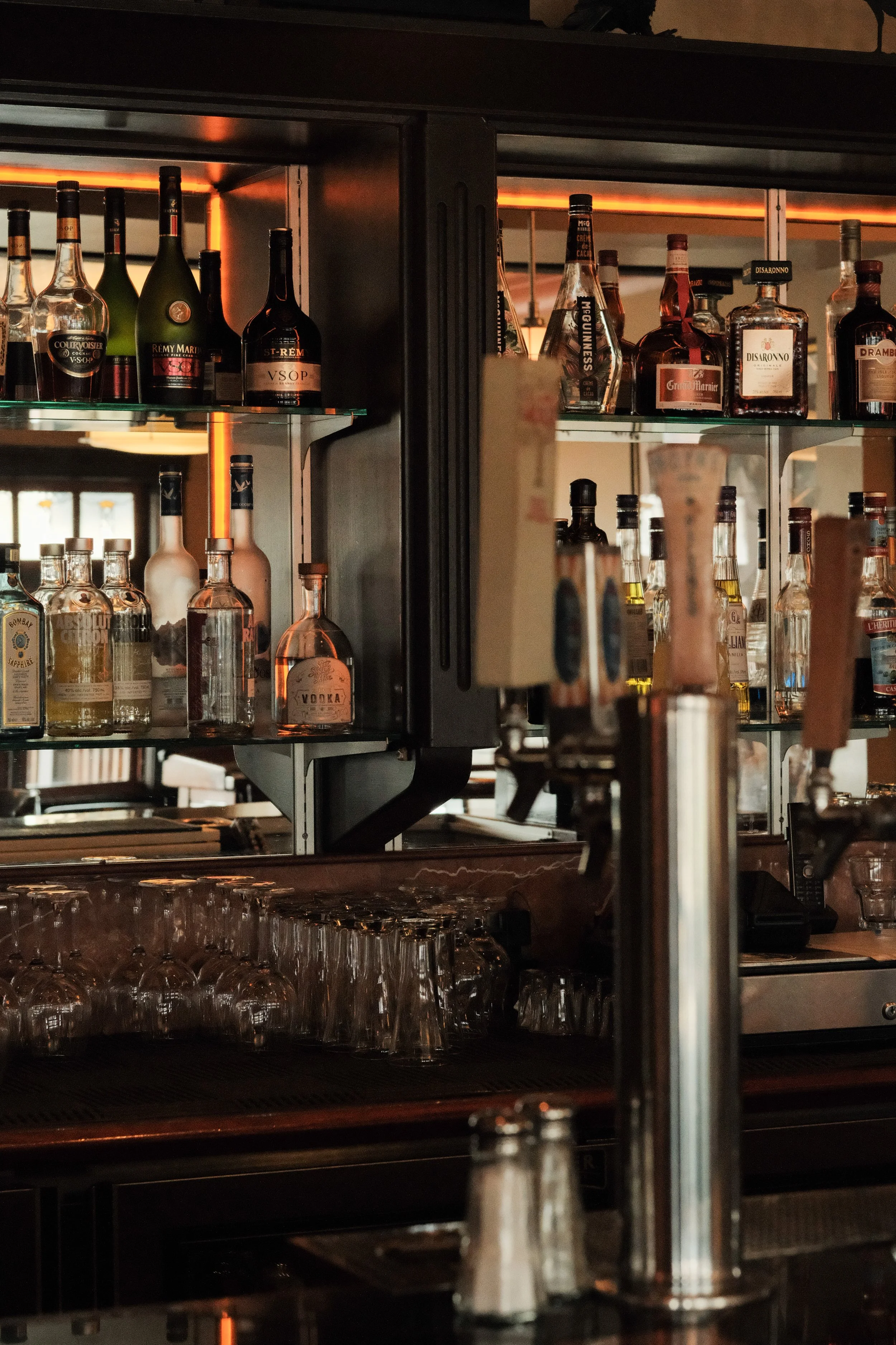 A bar shelf filled with various bottles of alcohol, including spirits and liquors. The bottles are arranged on glass shelves inside a black metal frame, with some reflective surfaces and warm lighting.