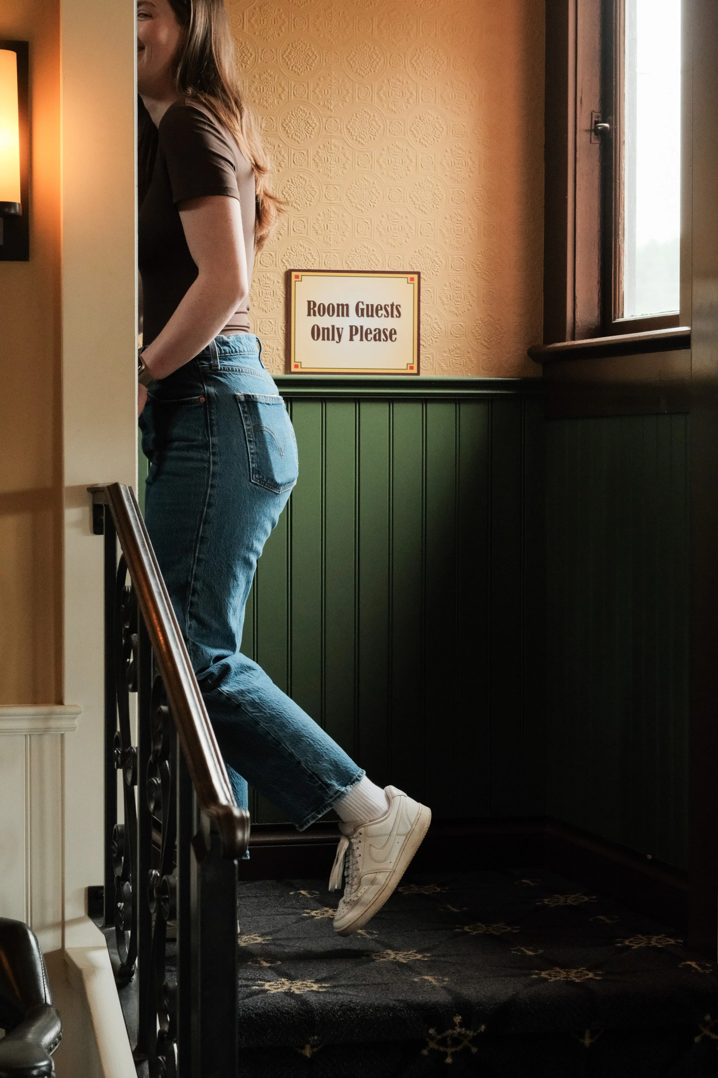 A person standing on a staircase with only the right side of their body visible, wearing a brown t-shirt, blue jeans, and white sneakers, beside a window with a sign that says "Room Guests Only Please" in a cozy, warmly-lit room.