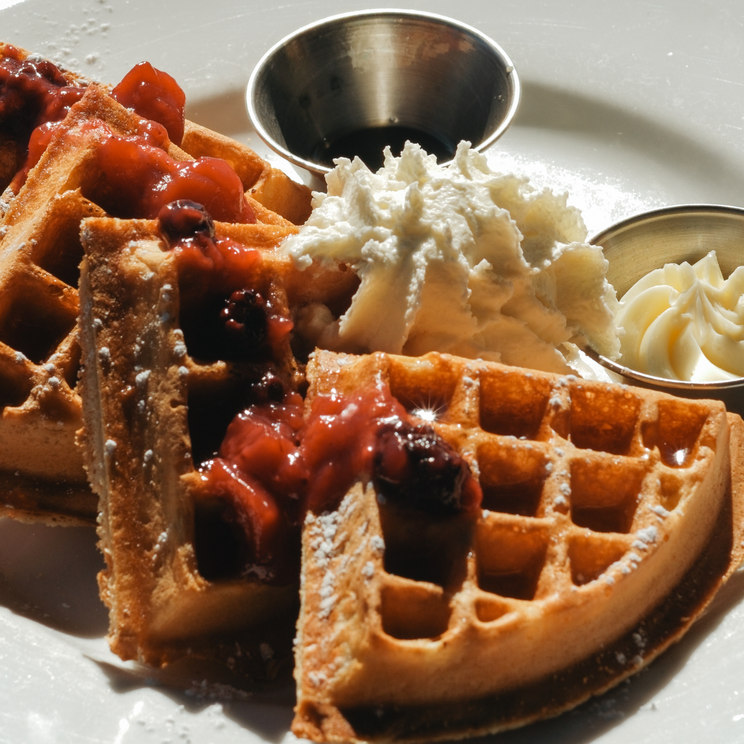 Two Belgian waffles topped with mixed berries, powdered sugar, served with whipped cream, syrup, and butter.