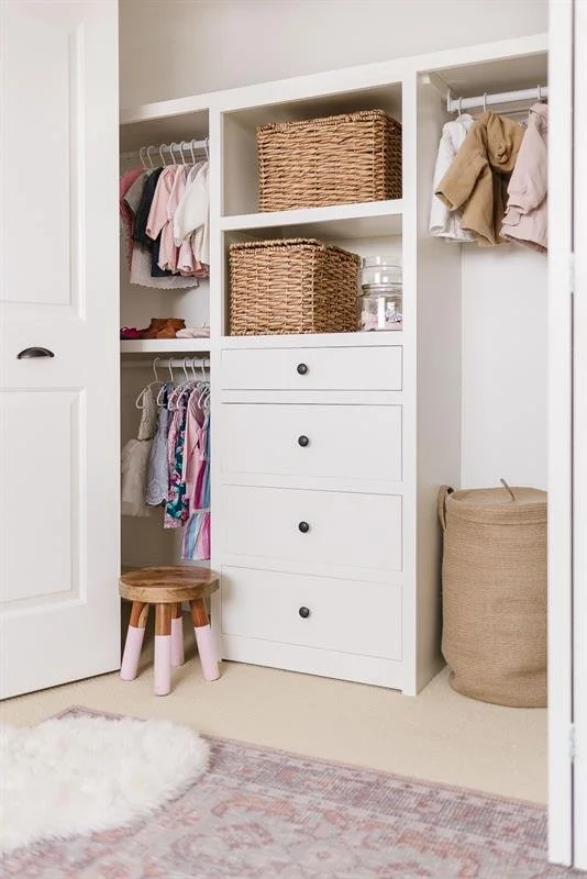 A white closet organizer with open shelves, drawers, and hanging rods filled with children's clothes, a woven basket, a glass jar, a small wooden stool, a beige laundry basket, and a soft pink and white rug on the floor.