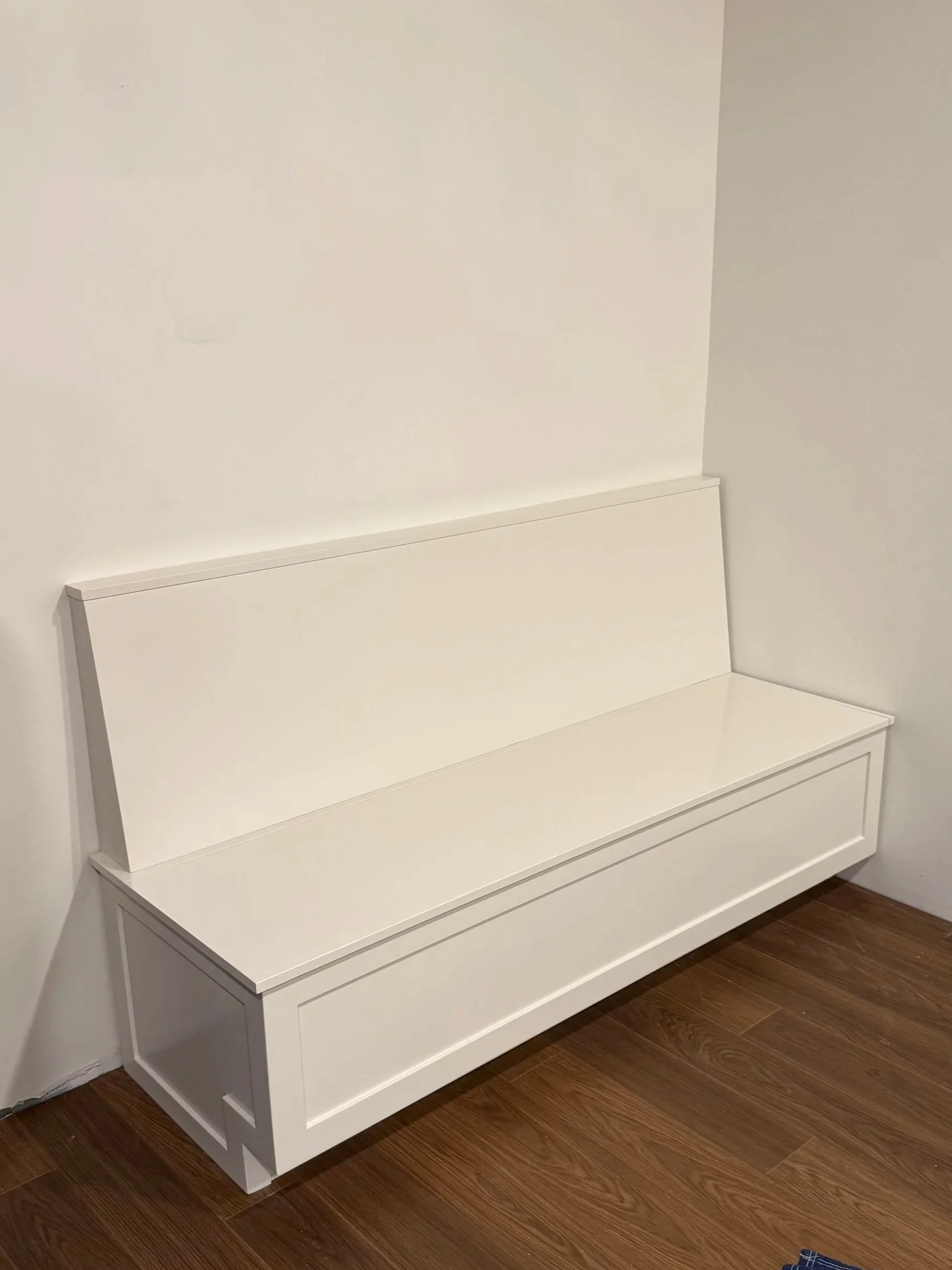 A white wooden bench with a backrest and a drawer underneath, placed against a white wall on a dark wood floor.