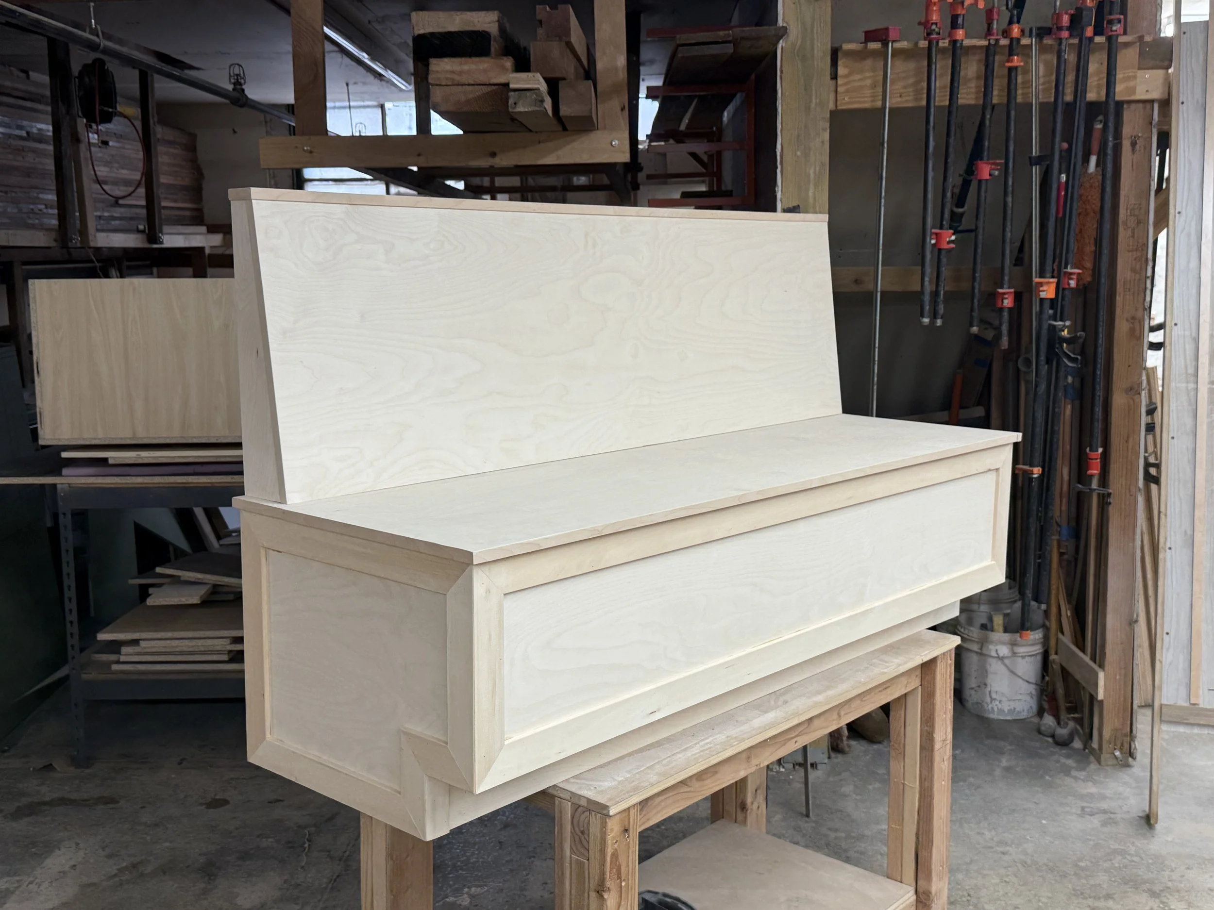 Unfinished wooden furniture piece in a workshop, with clamps on the right side and wooden shelves in the background.