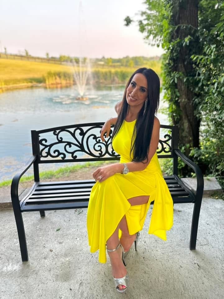 A woman in a bright yellow dress sitting on a black metal bench near a pond with a fountain, surrounded by greenery and trees.