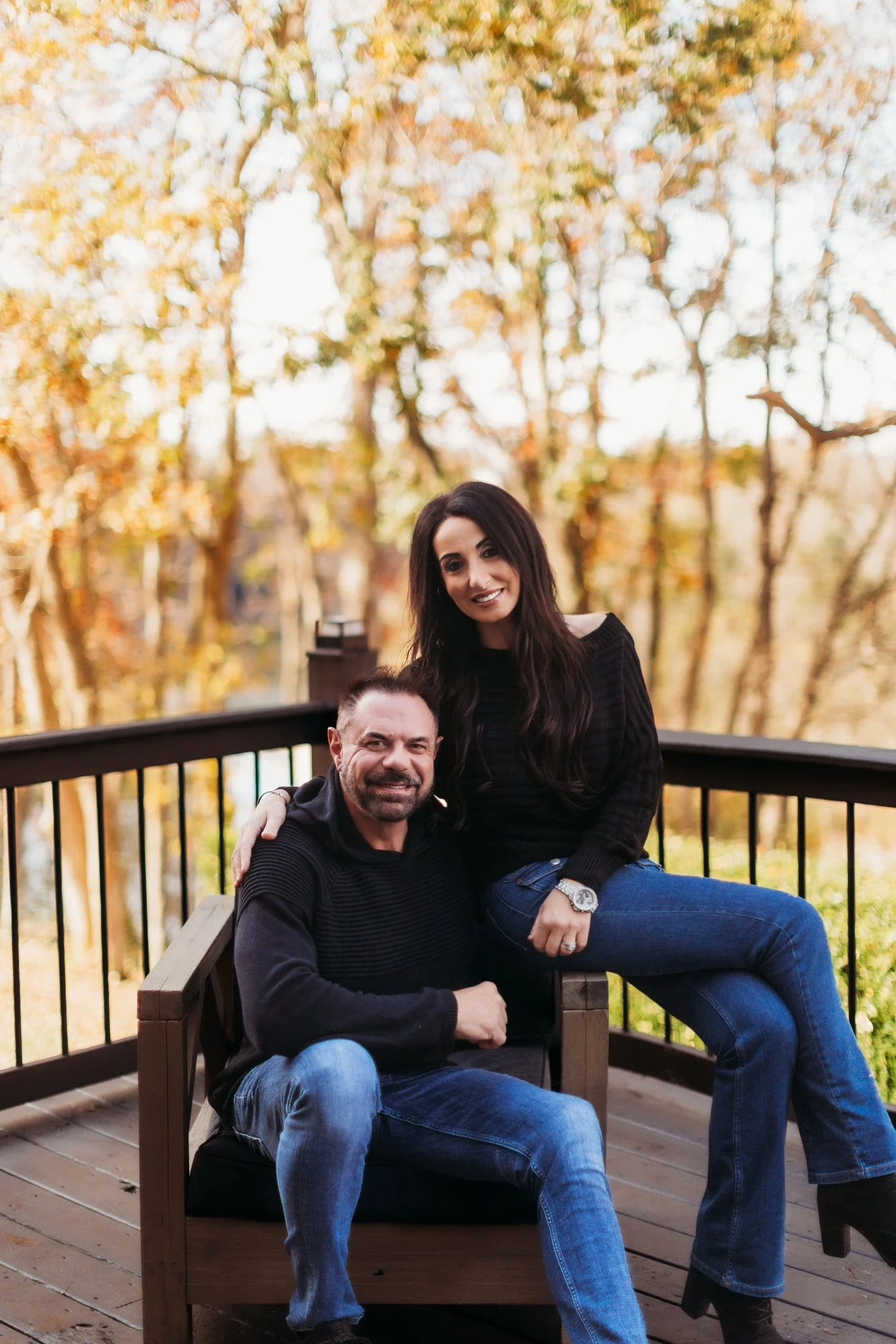 A smiling couple sitting on a wooden bench outdoors during fall, with trees and orange leaves in the background.