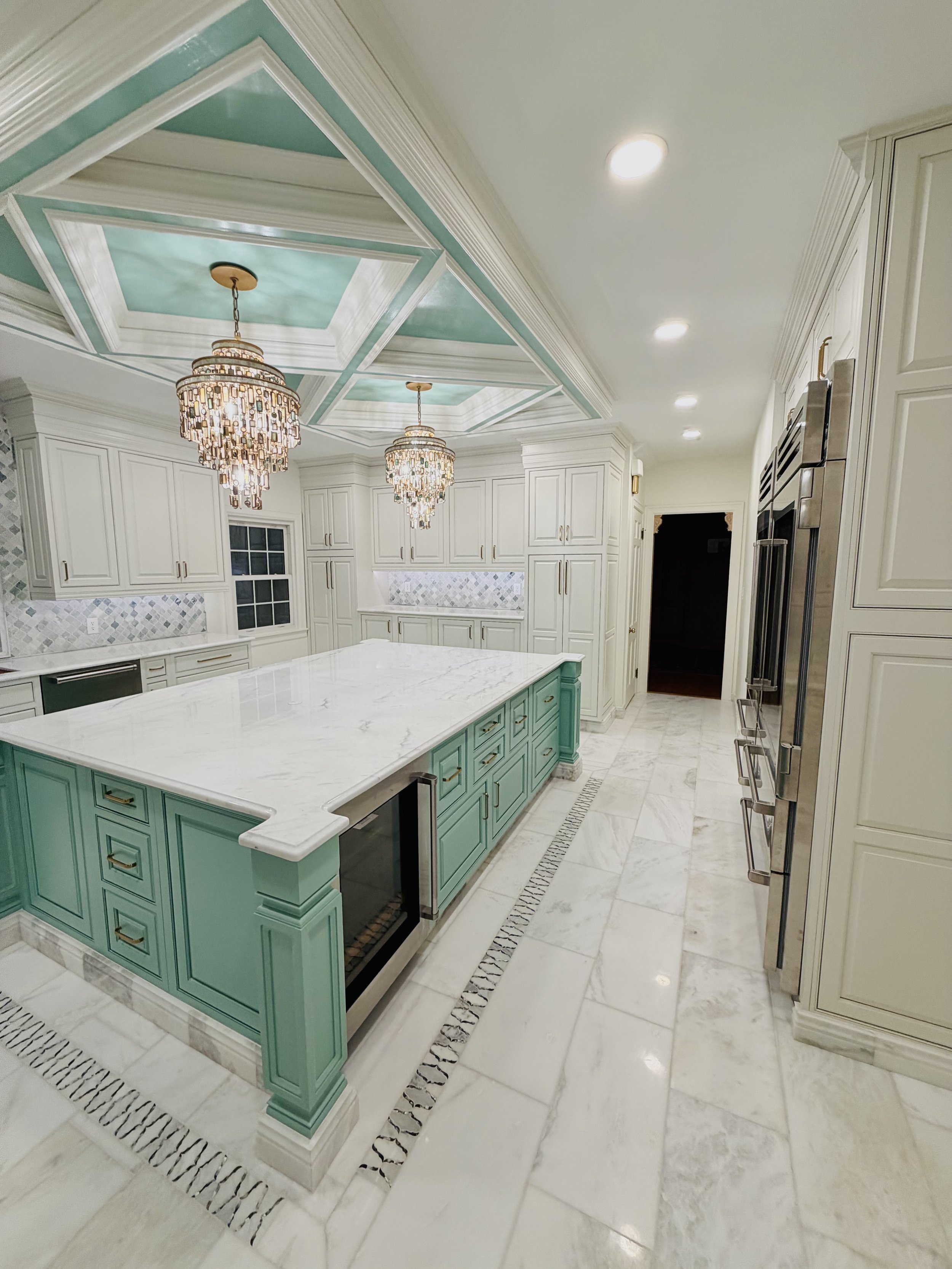 A spacious kitchen with white cabinets, marble countertops, and a central island painted in turquoise. Two elegant chandeliers hang from a ceiling with turquoise painted panels. Recessed lights illuminate the room, and there are stainless steel appliances including a double oven and a large refrigerator. The floor is made of white marble with a decorative border.