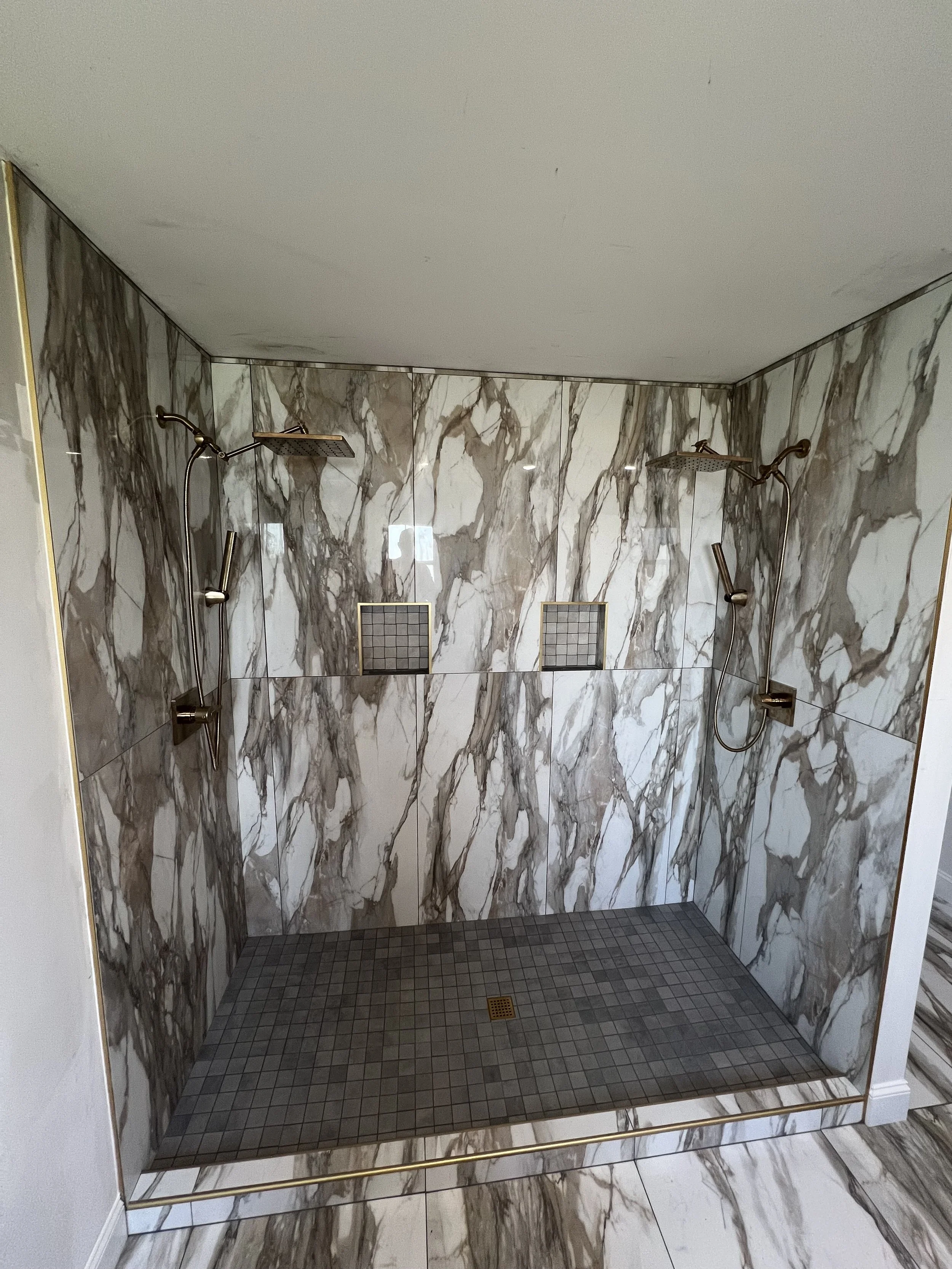 A modern, marble-tiled walk-in shower with two shower heads, handheld and overhead, and two small tiled niches on the back wall for toiletries. The shower has a gold railing around the entrance.