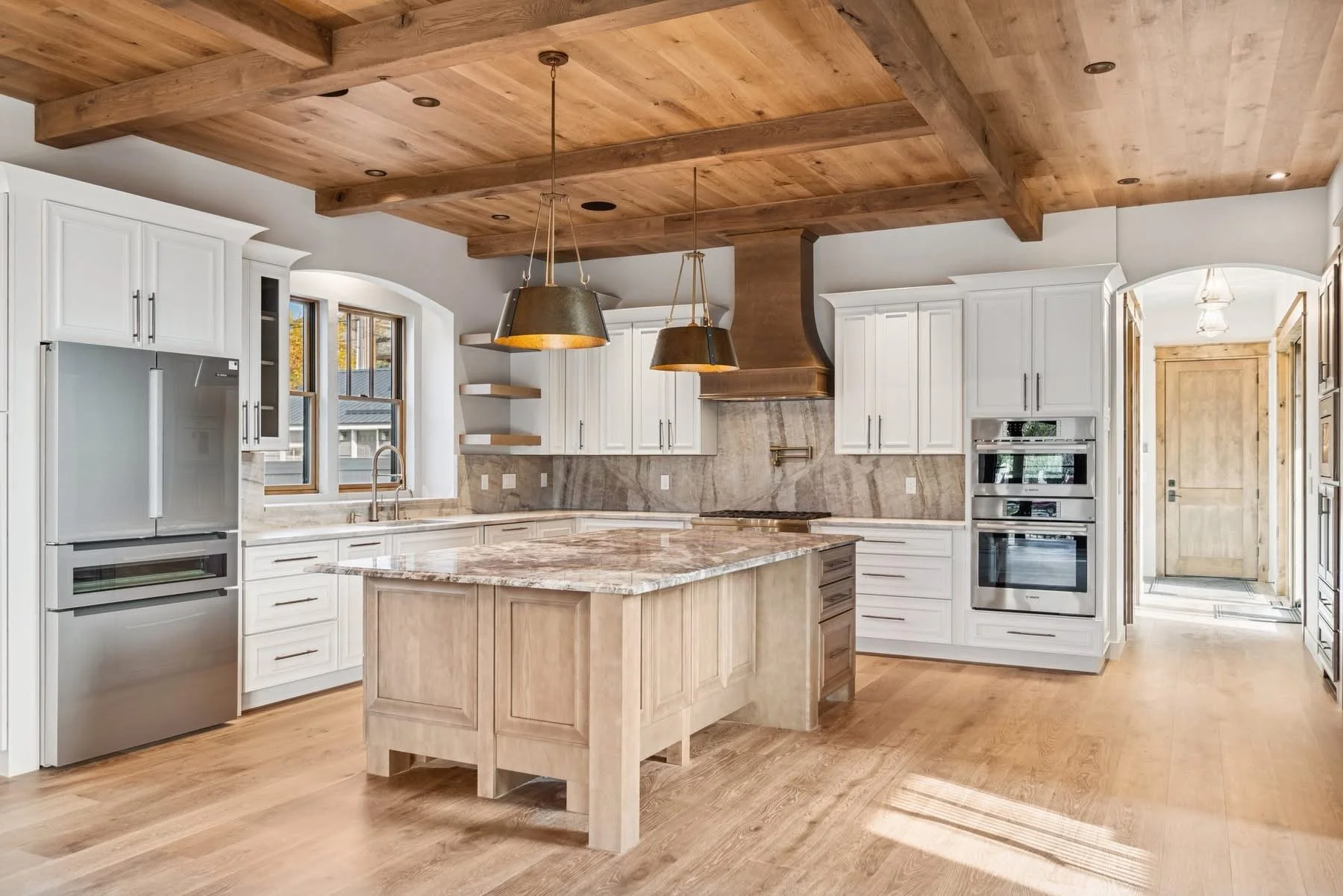 Modern kitchen with white cabinets, stainless steel appliances, a central island with marble countertop, wooden ceiling with beams, and hardwood floors.