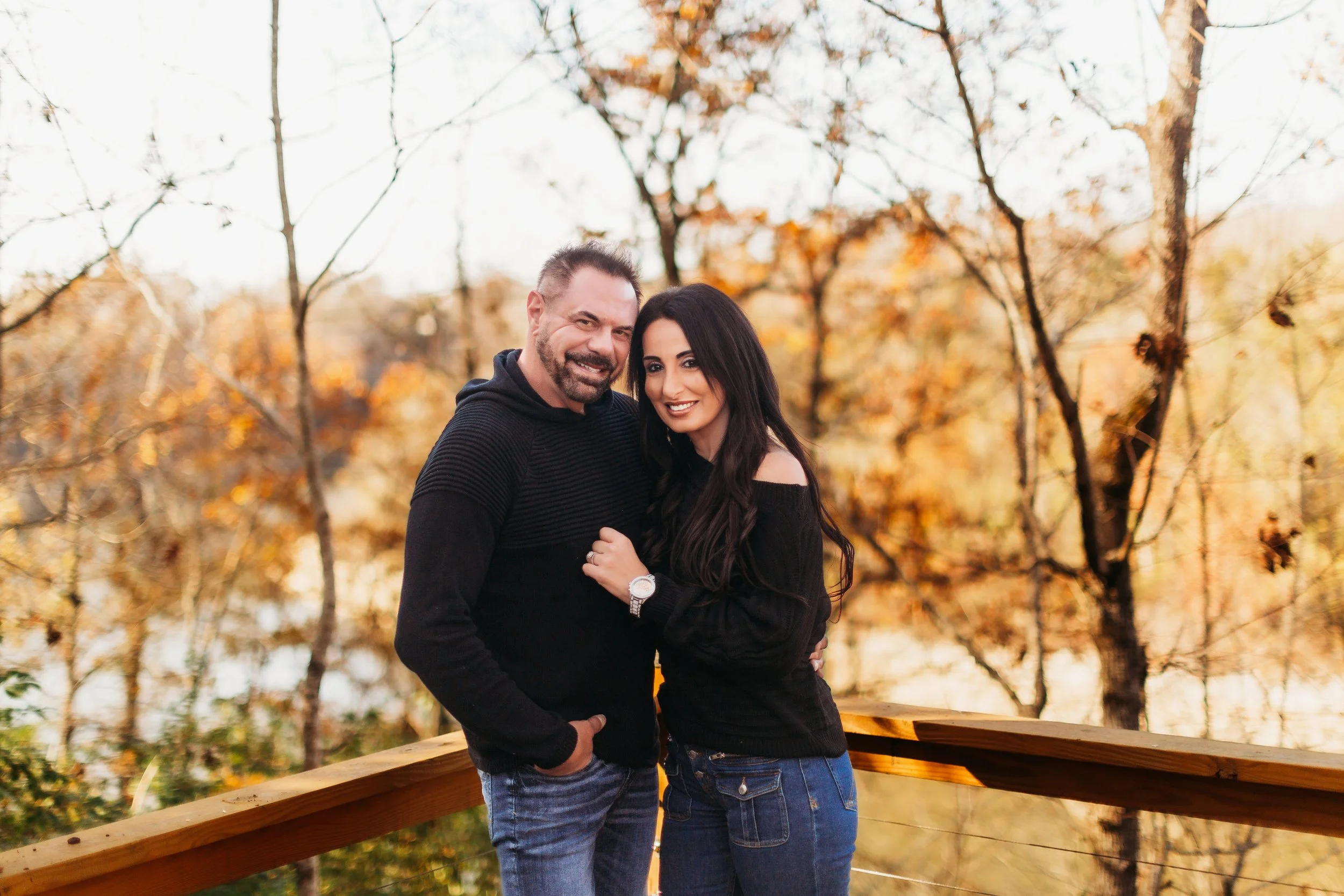A smiling man and woman stand close together on a wooden bridge with a fall landscape of trees with orange and yellow leaves in the background during the daytime.